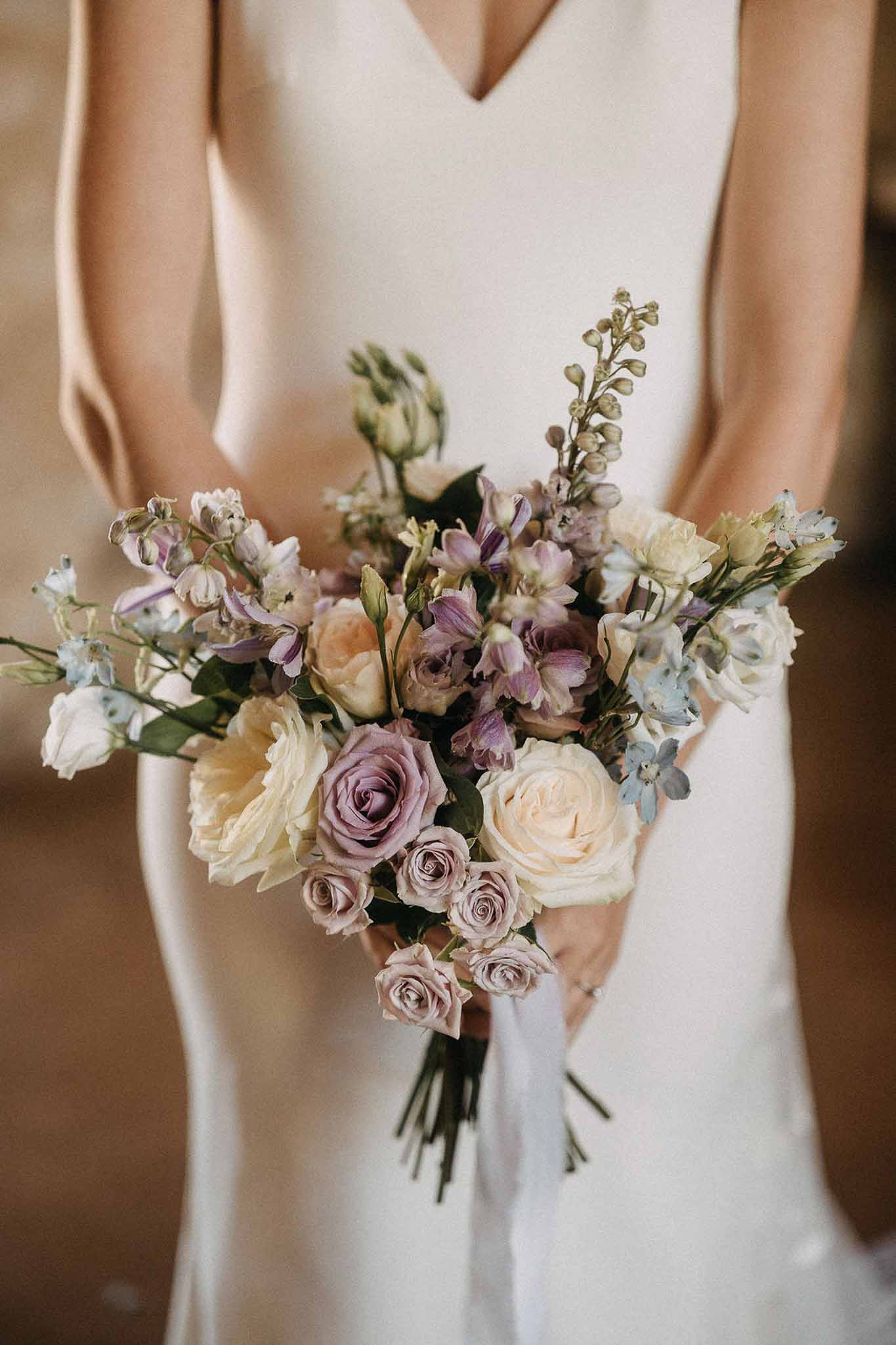 Garden-style bouquet of mauve roses, blue delphinium, and peach spray roses with trailing white ribbon