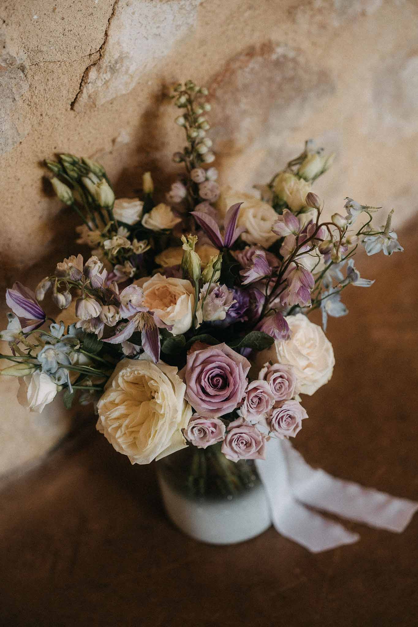 Mauve and dusty rose bouquet with purple clematis, blue delphinium, and white silk ribbon on plaster wall