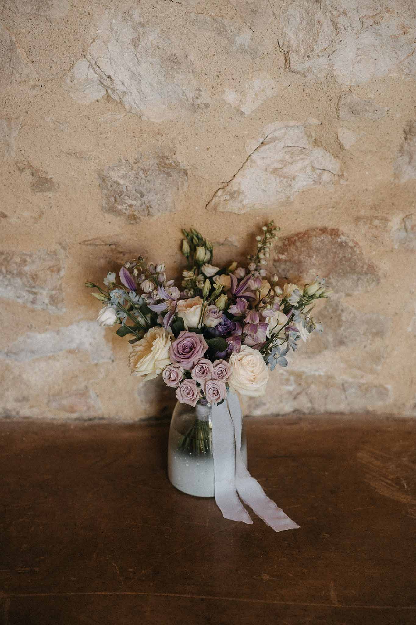 Loose garden bouquet of mauve roses, cream roses, and blue forget-me-nots in glass jar with silk ribbon