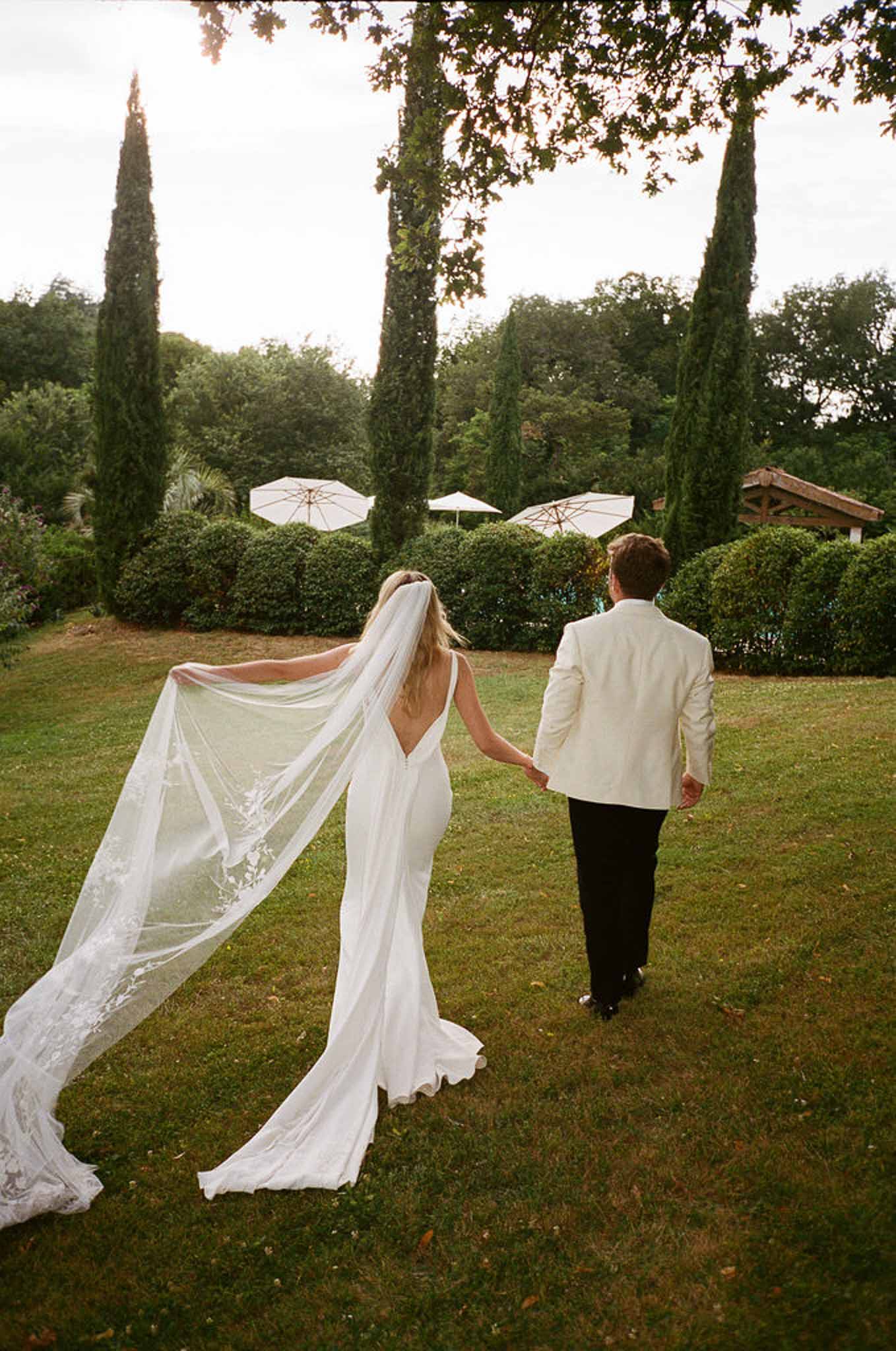 Couple walking from behind across lawn with lace-edged cathedral veil fanning out beside white parasol garden