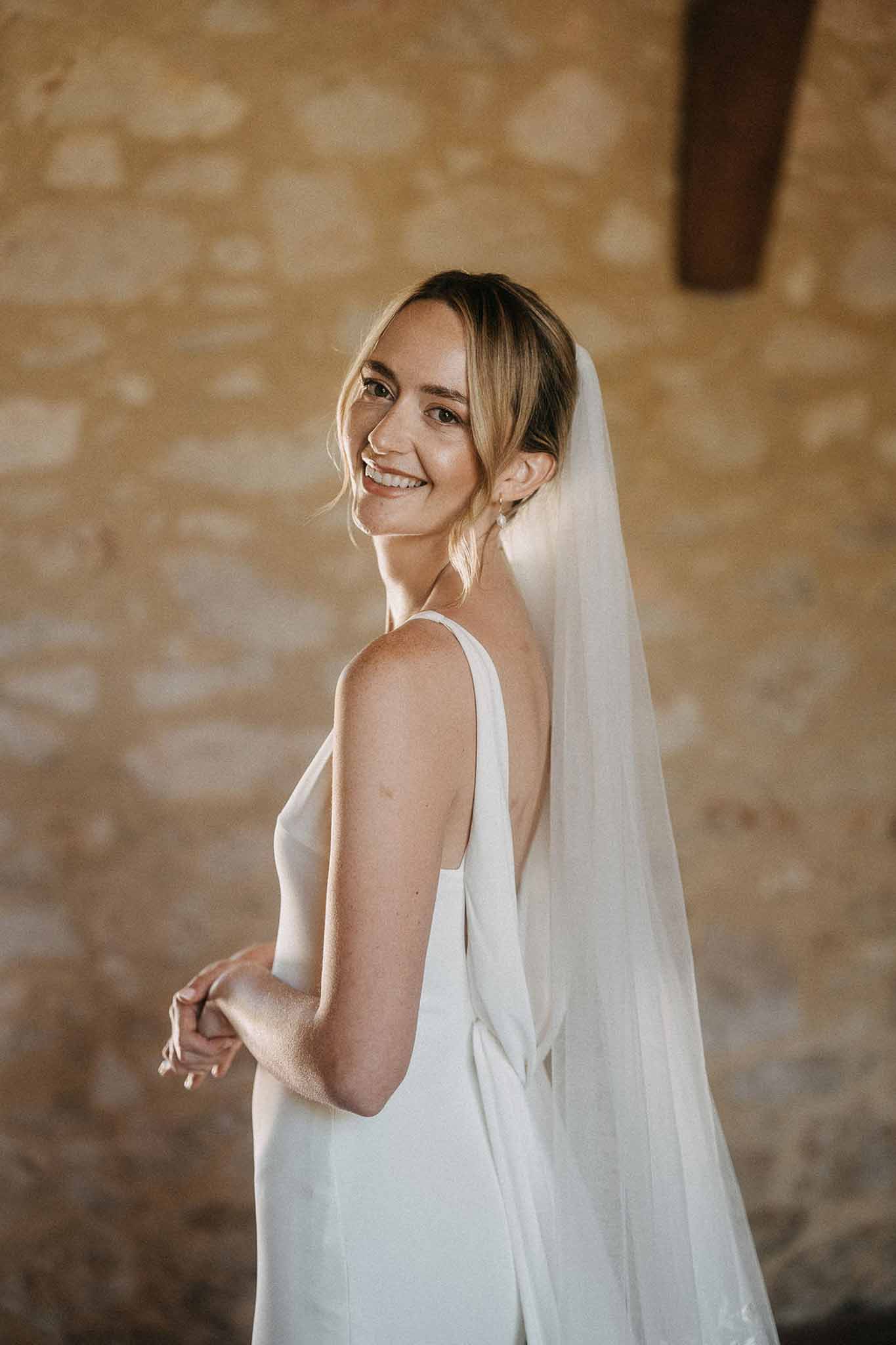 Bride glancing over shoulder showing low-back slip dress and veil against exposed stone wall