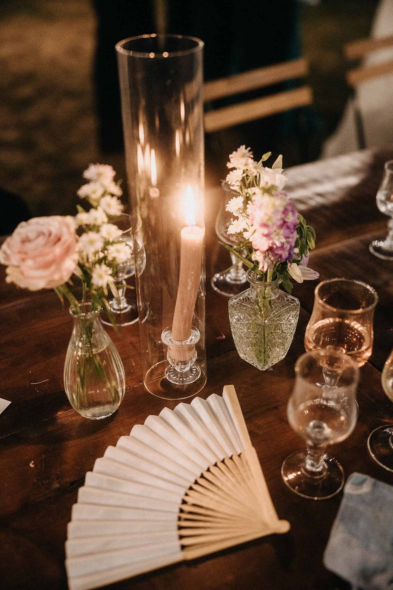 Reception table centerpiece with blush roses and purple blooms in mismatched glass vases beside a lit taper candle