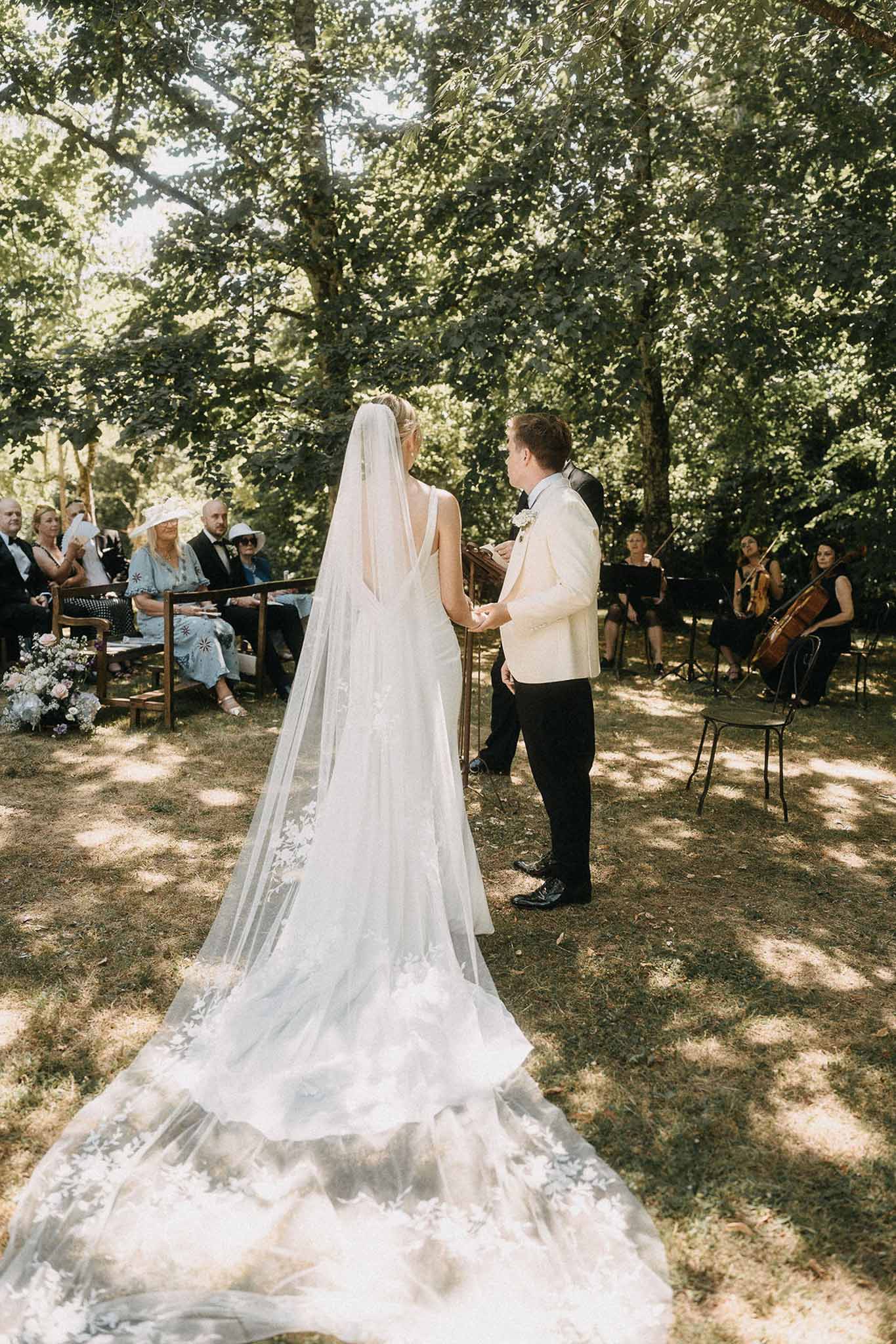 Garden wedding ceremony with couple exchanging vows, bride in cathedral-length lace veil, string trio playing