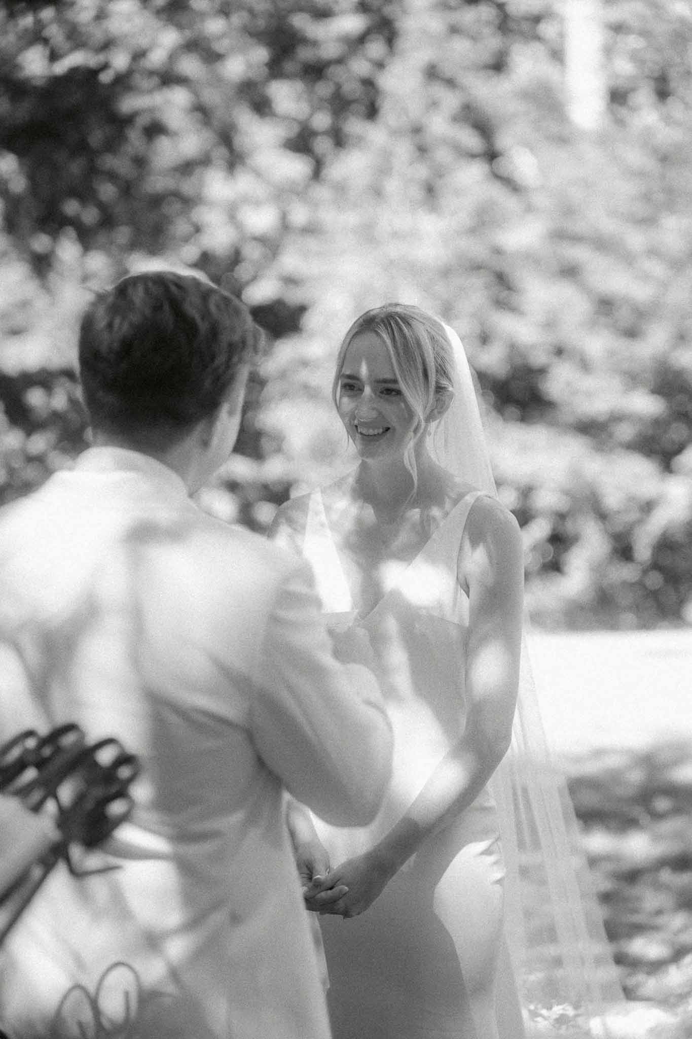 Black and white of smiling bride in fitted gown and veil holding hands with groom during outdoor ceremony