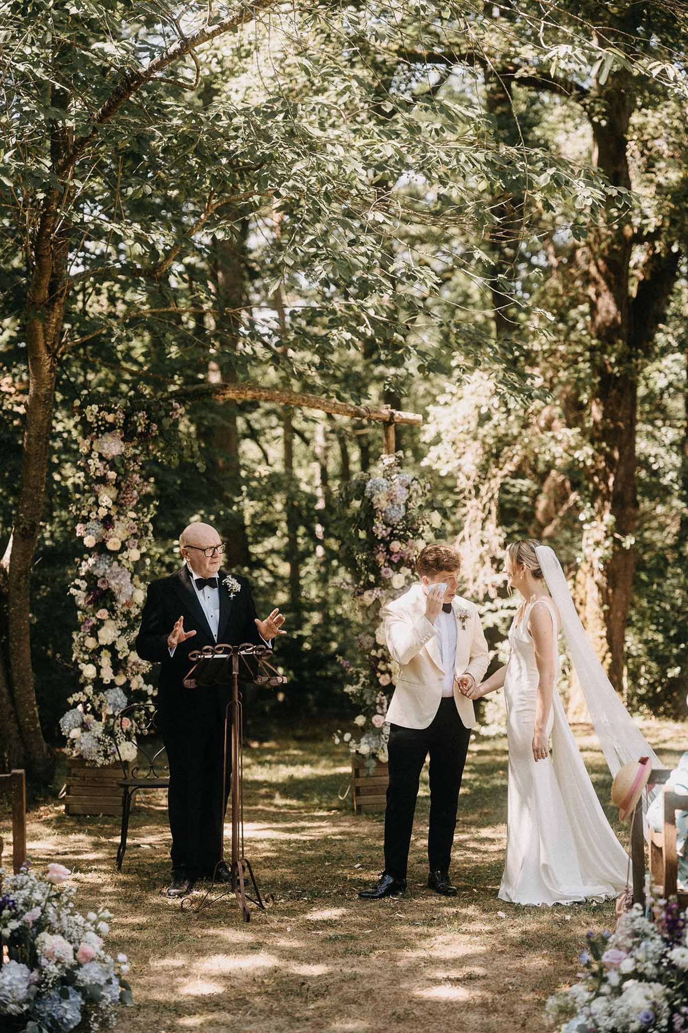 Emotional groom wiping eye at woodland altar with blush pink mauve and lavender floral columns bride in ivory satin