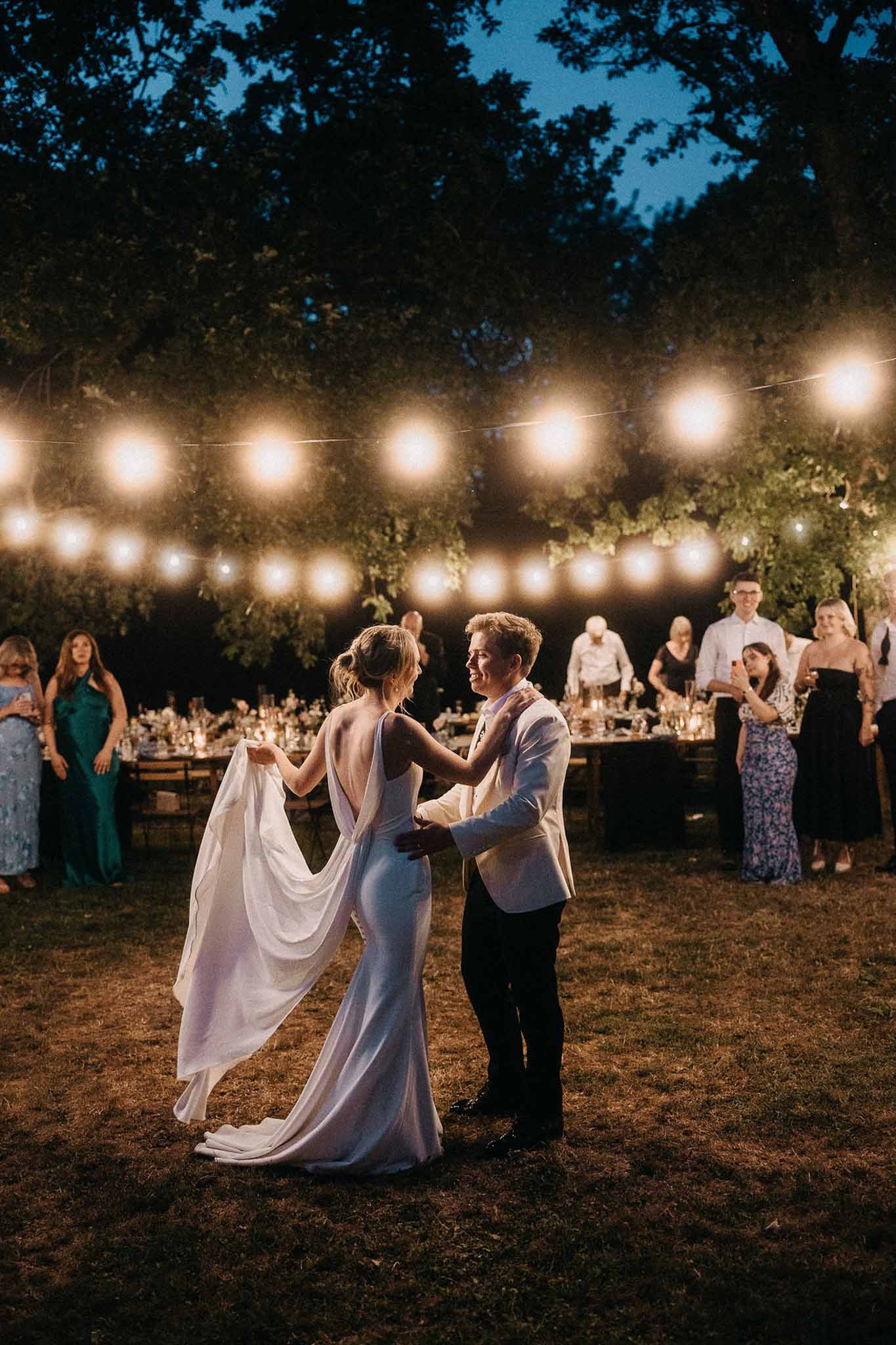 Couple sharing first dance on grass lawn at dusk under festoon lights with guests watching from dining tables