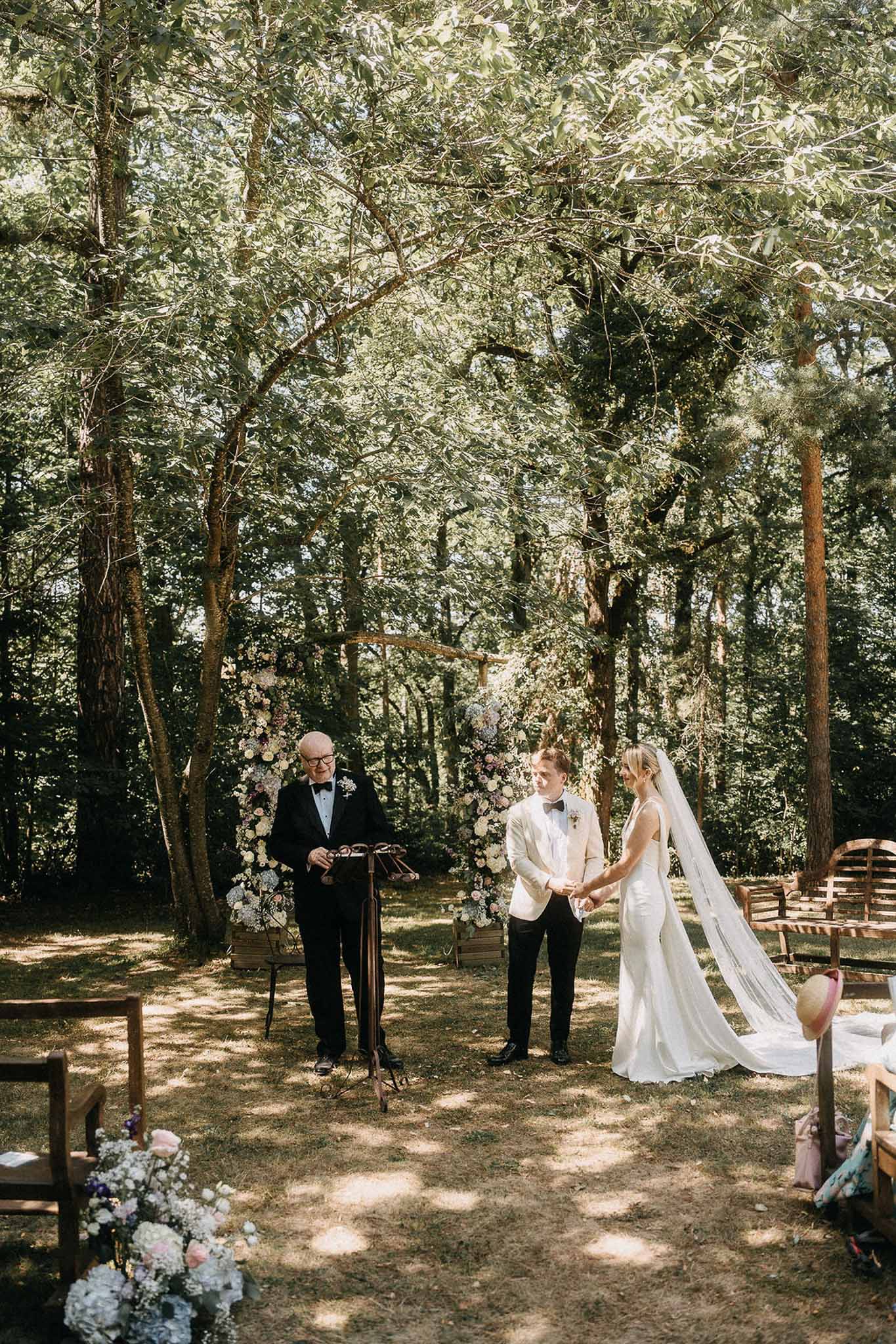 Outdoor woodland wedding ceremony with couple at altar flanked by pastel floral columns and seated guests