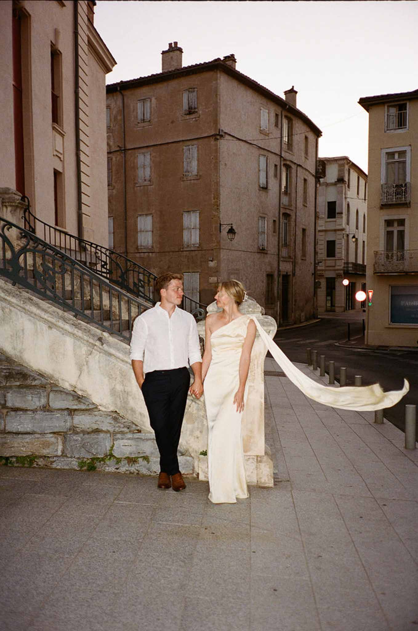 Couple walking hand in hand at dusk on French town square, bride in one-shoulder satin gown with train