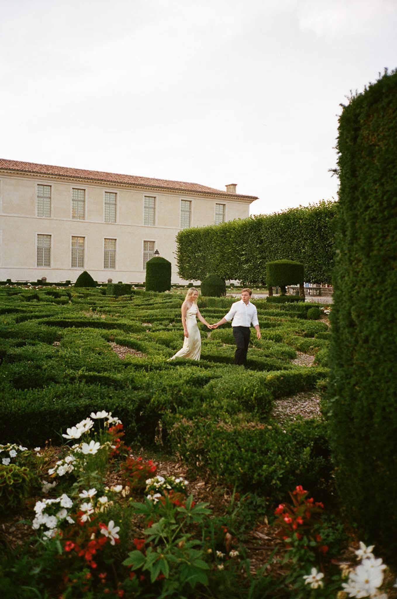 Couple walking hand-in-hand through formal French parterre garden with clipped boxwood hedges and topiary