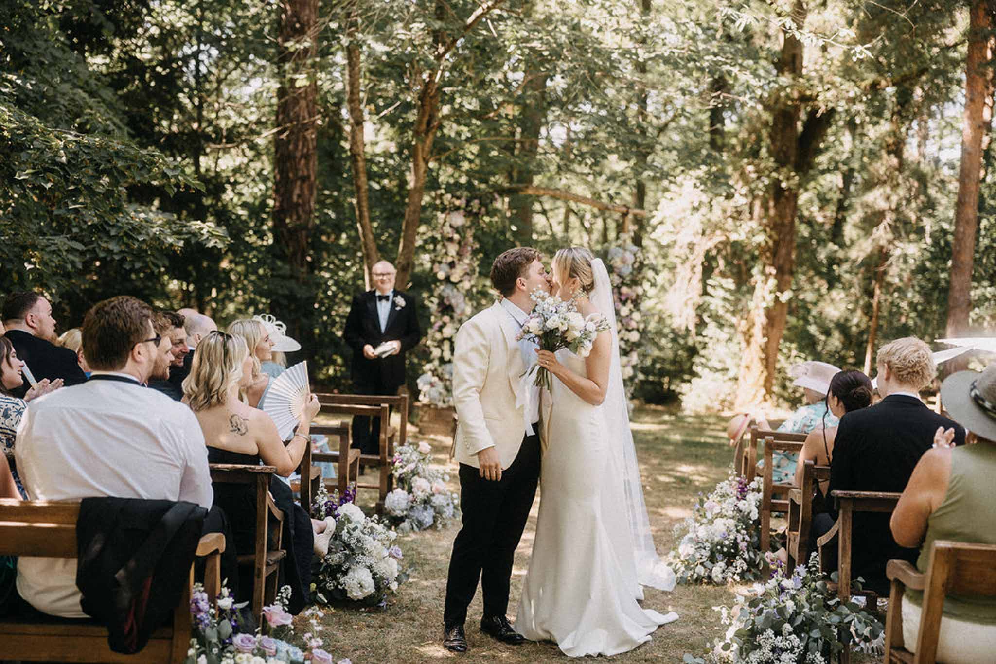 Couple shares first kiss during outdoor woodland ceremony with seated guests and pastel floral aisle arrangements
