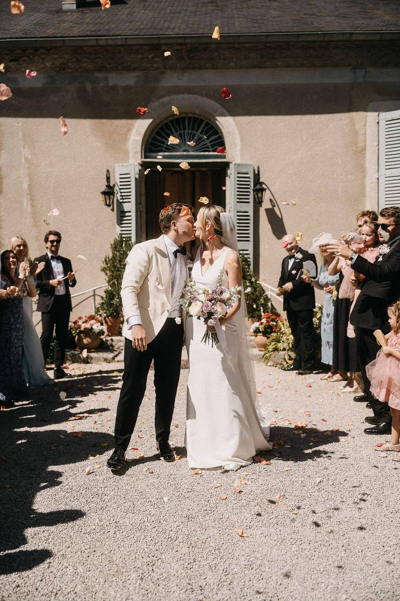 Couple kisses during petal toss ceremony exit as guests clap in gravel courtyard of French manor