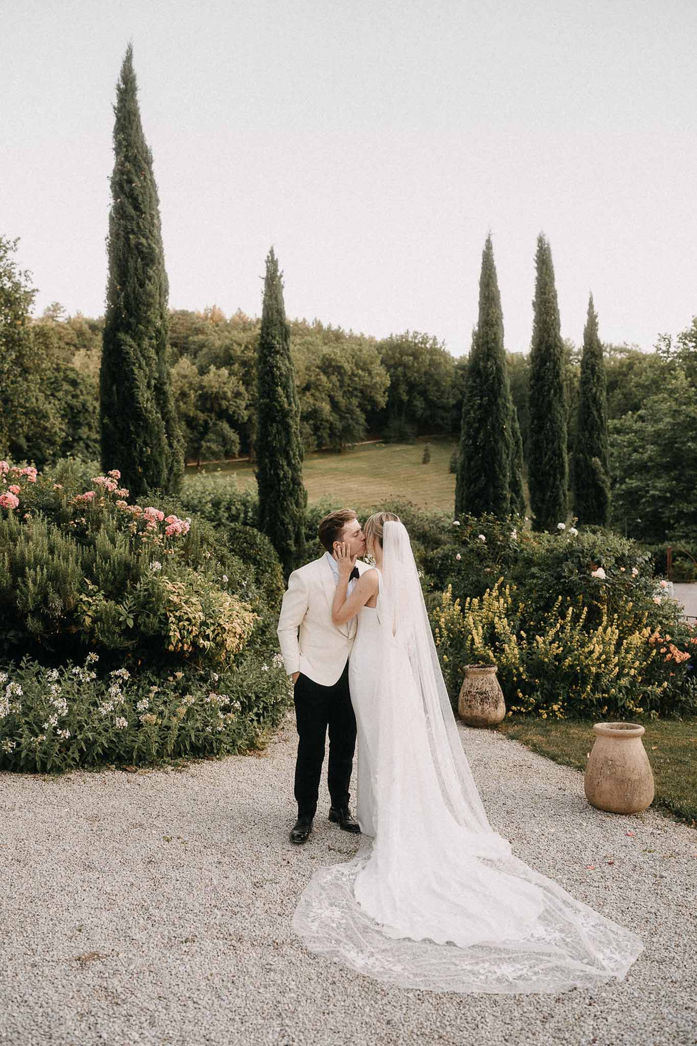 Couple kissing on gravel path with cathedral lace veil fanned out amid roses and cypress trees