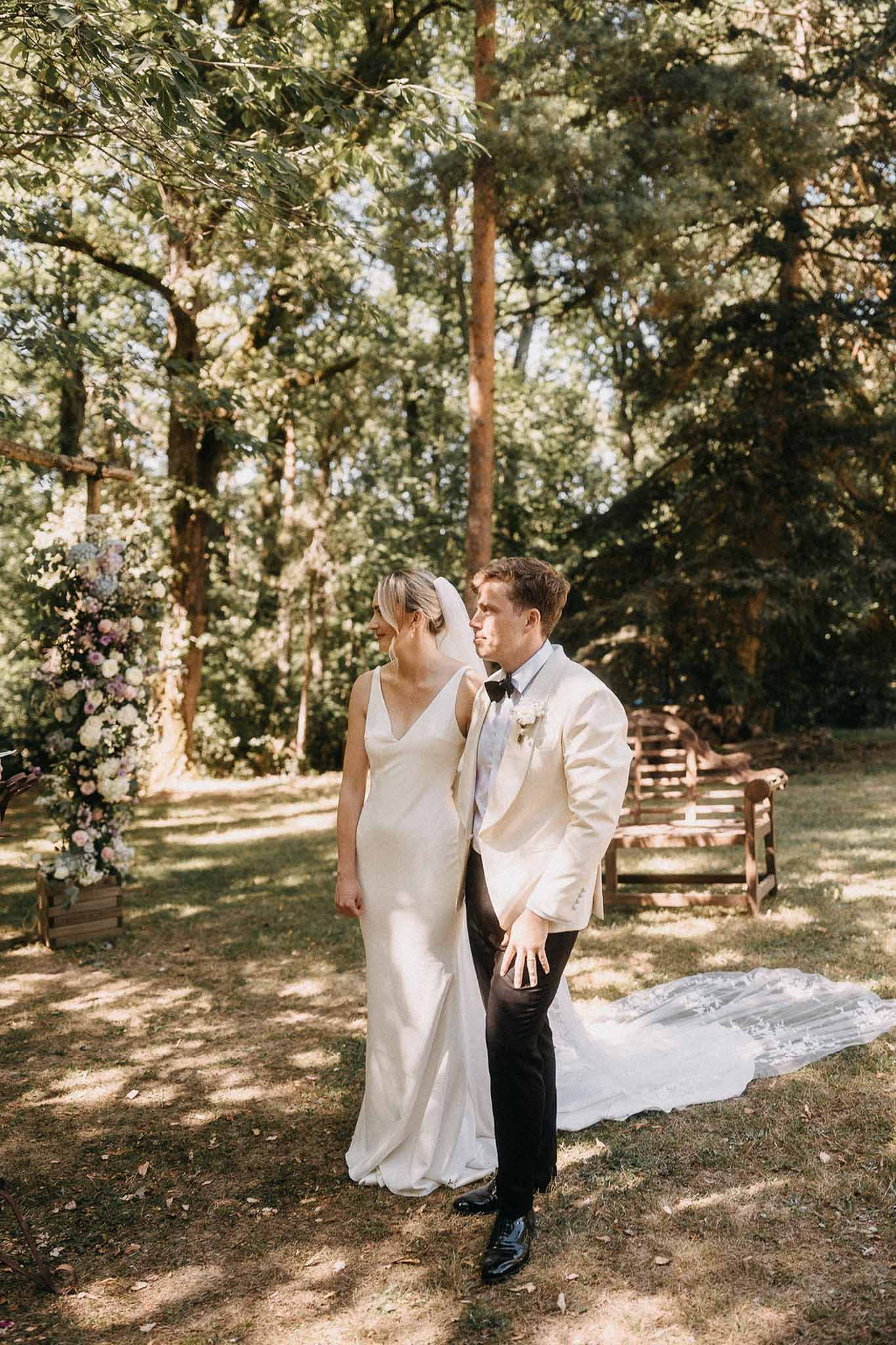 Couple standing before tall blush and lavender floral installation in wooded ceremony setting