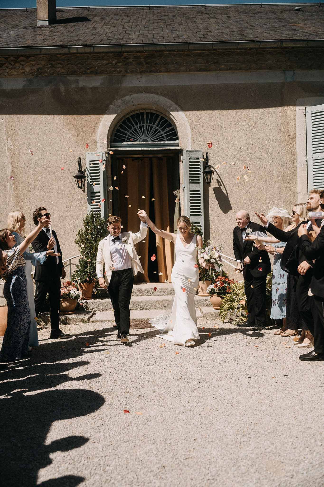 Bride in white satin gown and groom in cream blazer exit through pink petal confetti at manor courtyard