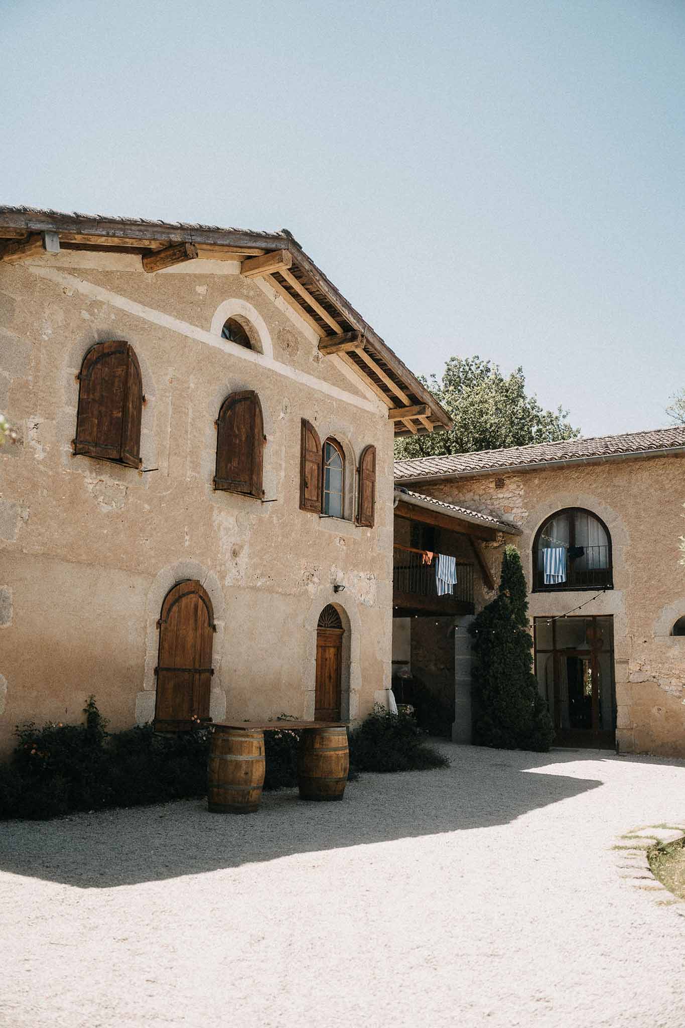 Rustic French farmhouse venue with beige stone walls, terracotta roof, wine barrels, and gravel courtyard