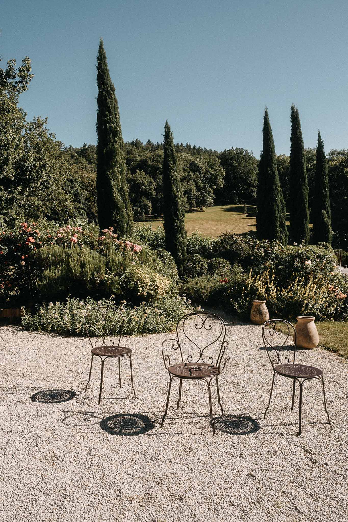 Wrought-iron bistro chairs and terracotta urns on gravel courtyard with cypress trees and flowering shrubs
