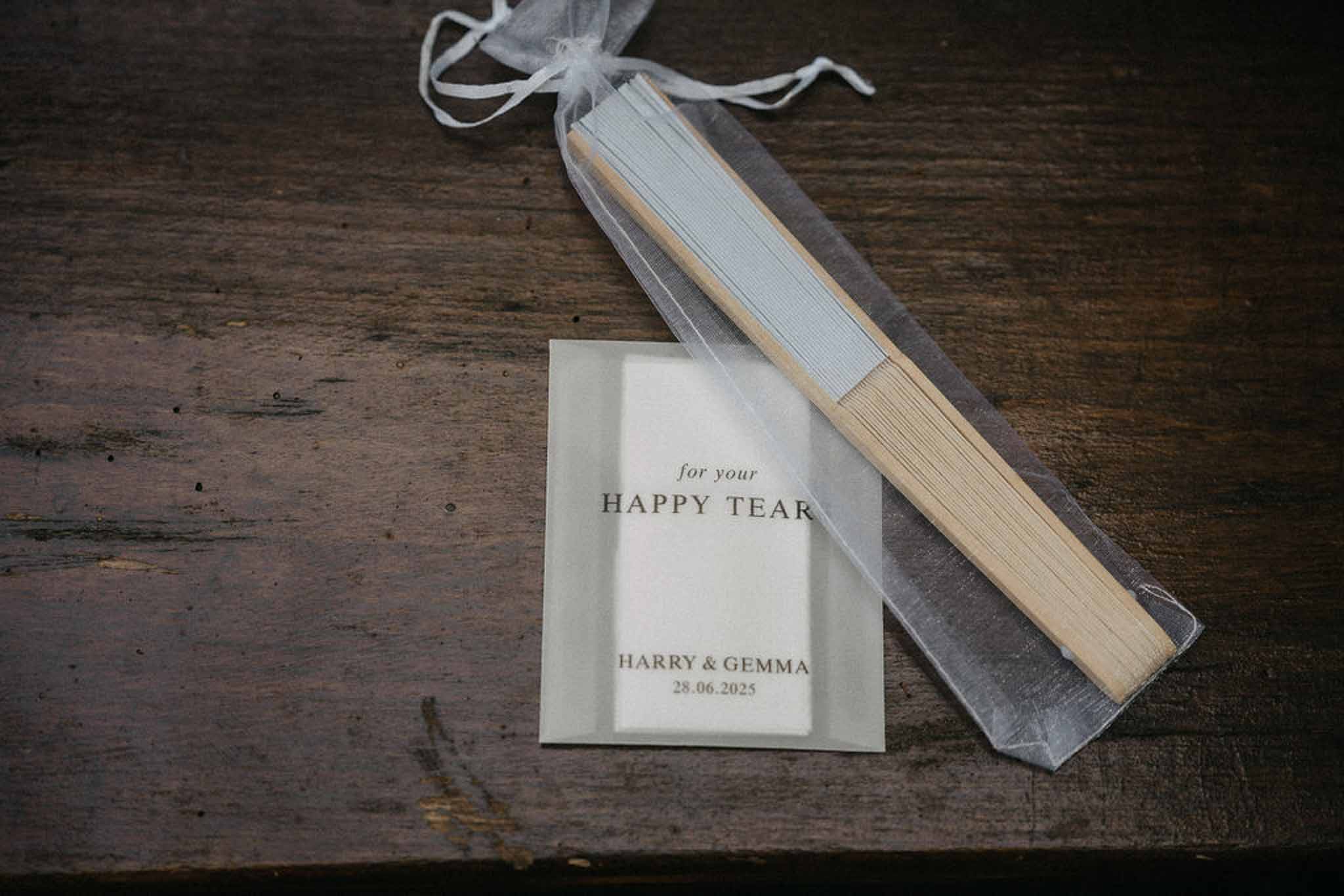 Wedding favour hand fan in organza bag with personalised happy tears card on dark wood surface