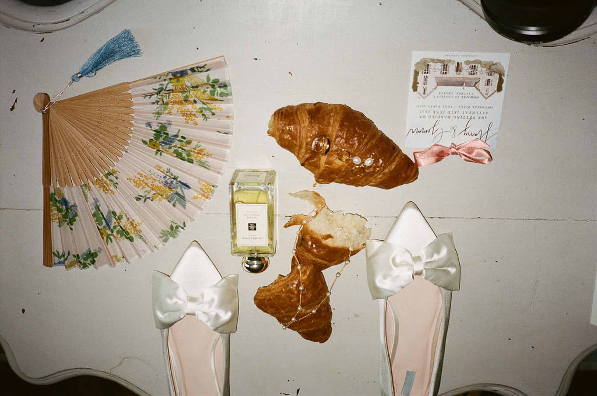 Bridal flat lay with floral fan, blush bow heels, perfume, croissant with rings, and chateau invitation