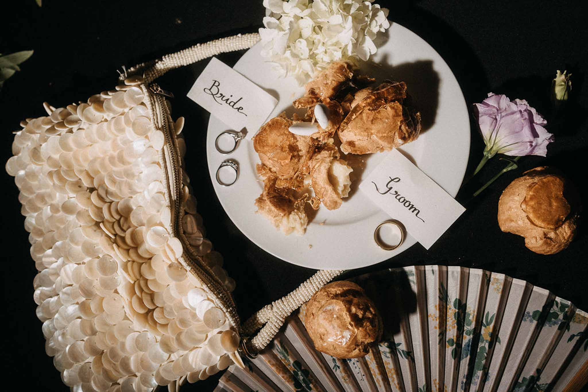 Flat lay of place cards, rings, sequin clutch, floral fan, and French choux pastries on dark surface