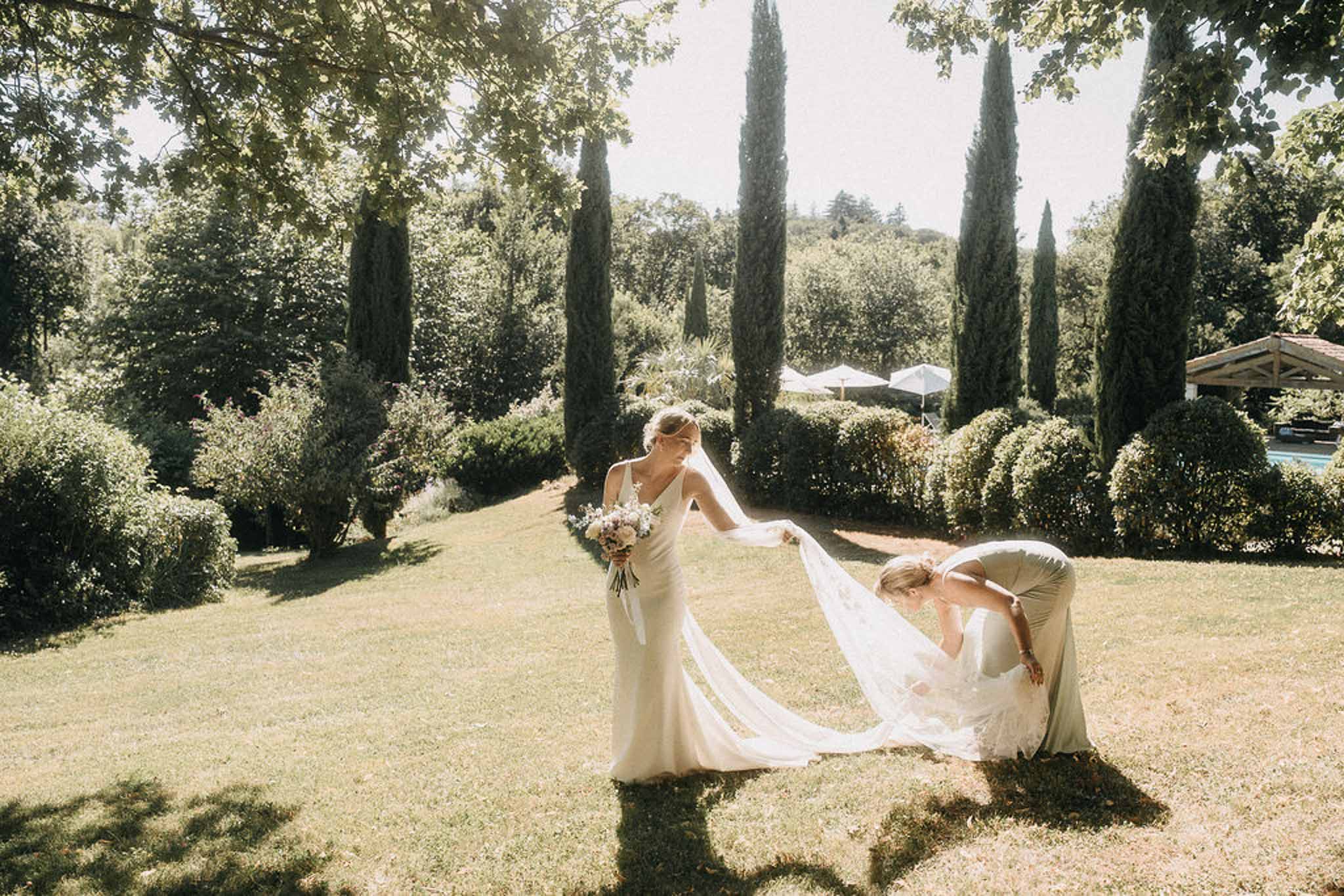 Bridesmaid in gold dress crouching to arrange bride's slip gown train in cypress-lined garden