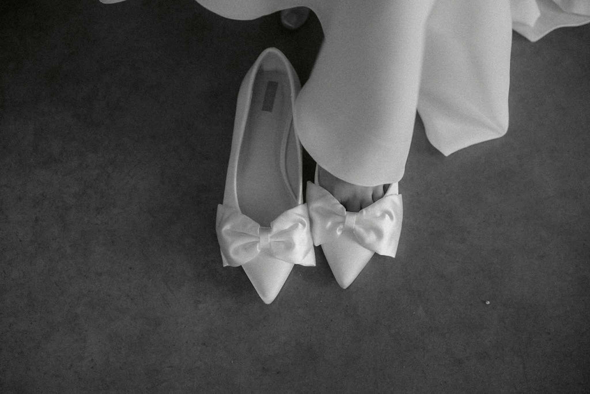 Black and white close-up pointed bridal flats with satin bow embellishments on dark floor with gown hem visible