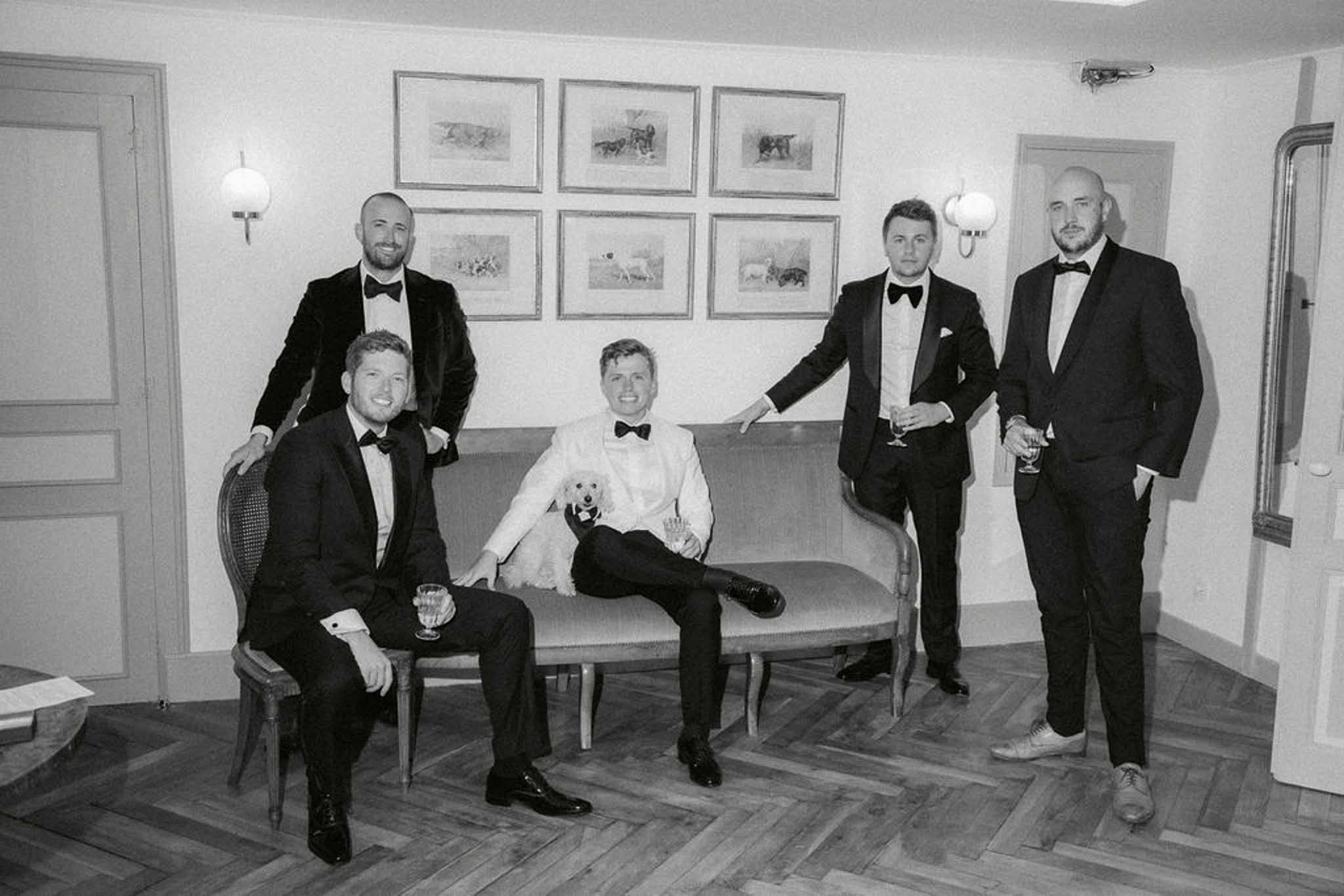 Groom in white dinner jacket with small dog and four groomsmen in tuxedos on settee in salon in B&W