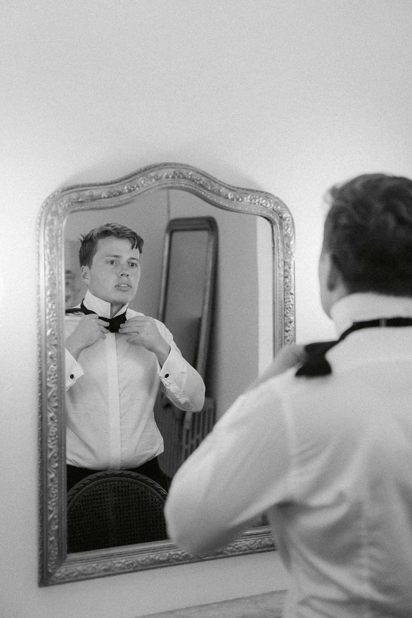 Groom adjusting bow tie in ornate carved mirror reflection wearing white shirt and suspenders in B&W