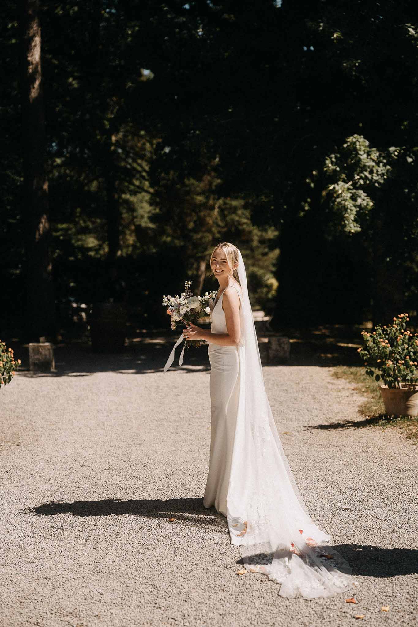 Bride looking over shoulder on chateau gravel driveway wearing ivory slip gown with cathedral veil and white bouquet