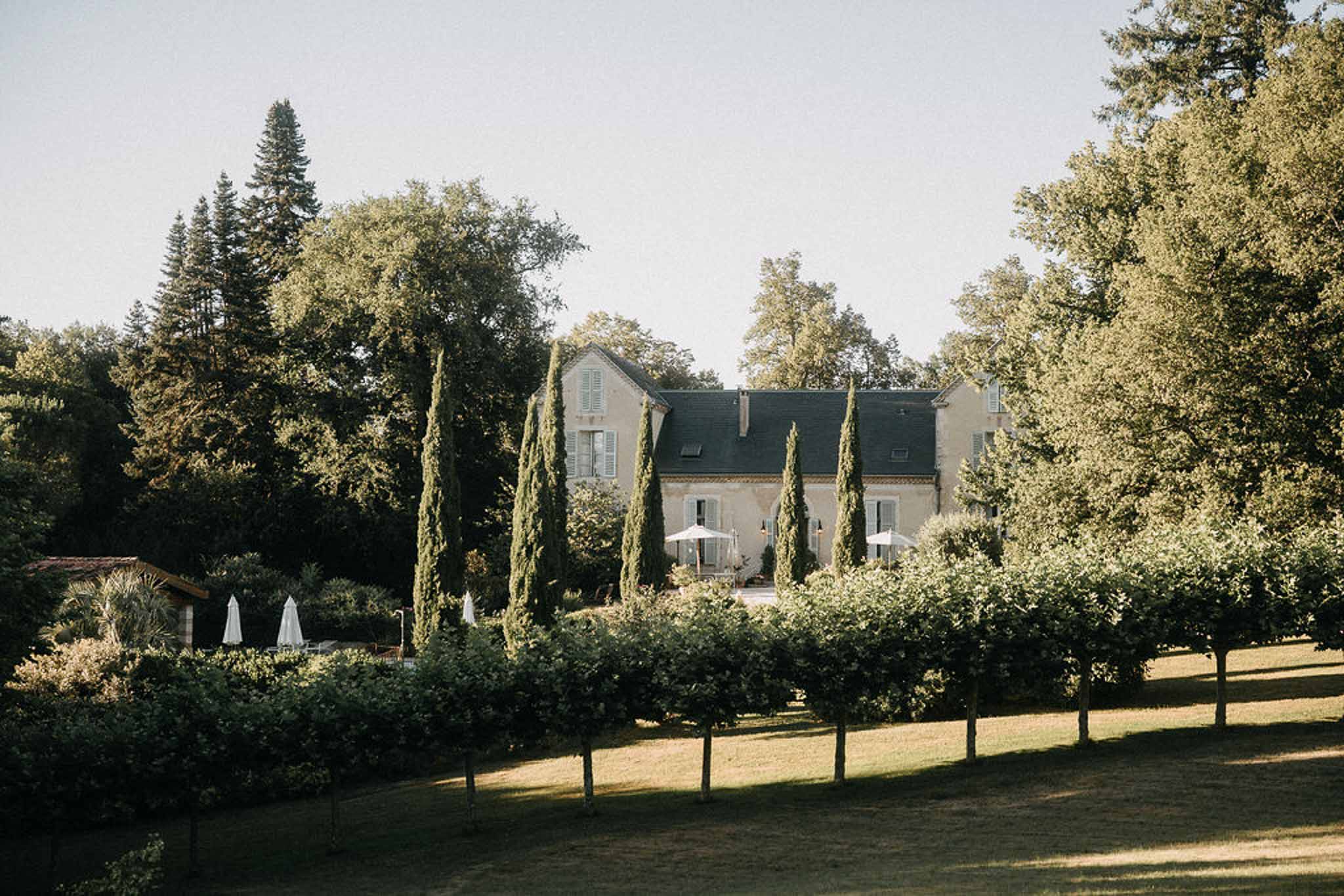 Cream manor house with slate roof and blue shutters behind cypress row with pool outbuilding at left