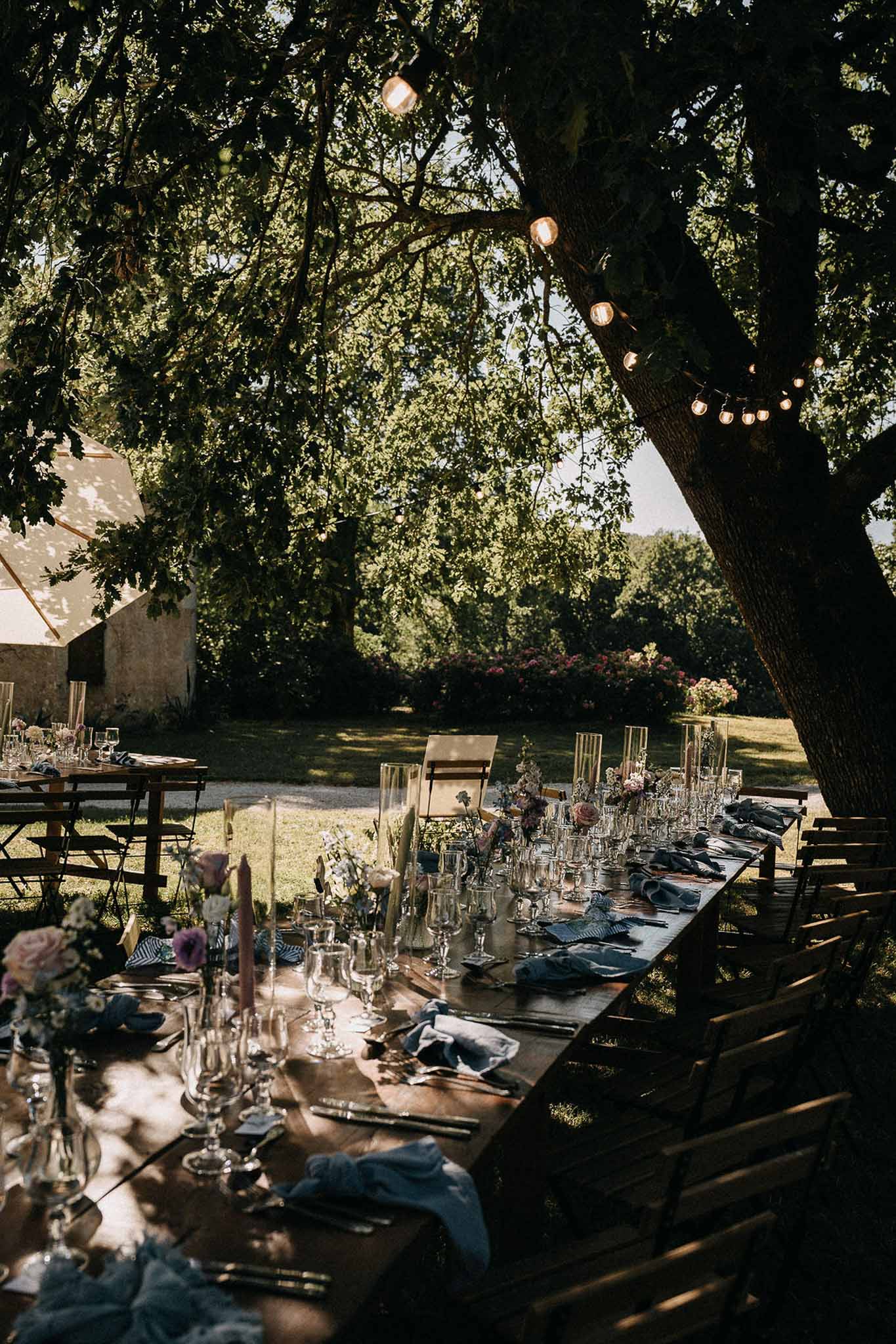 Outdoor reception table under trees with dusty pink candles, blush roses, purple blooms, and globe string lights