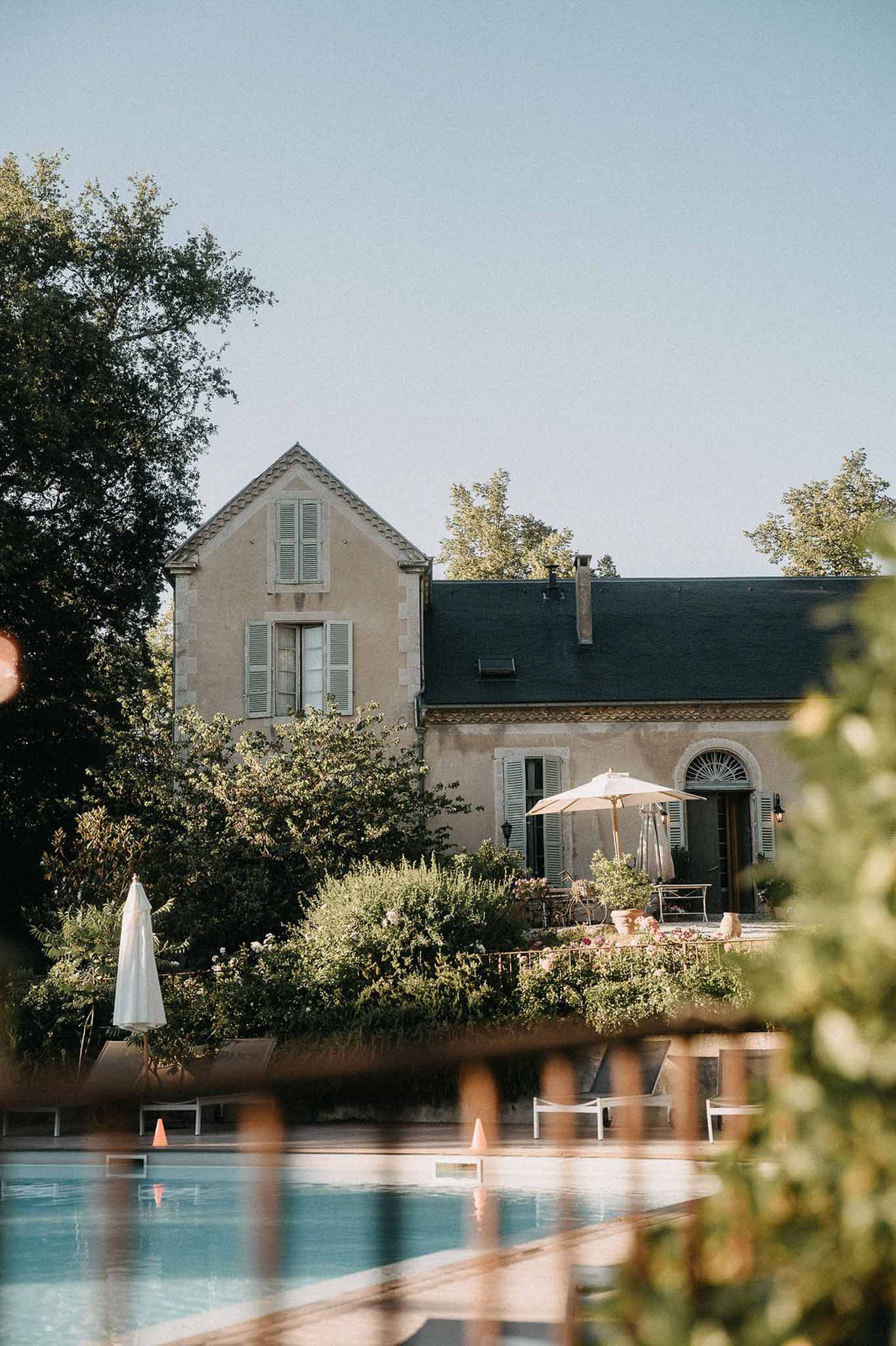 French manor house viewed across swimming pool with cream parasol, white loungers, and slate roof