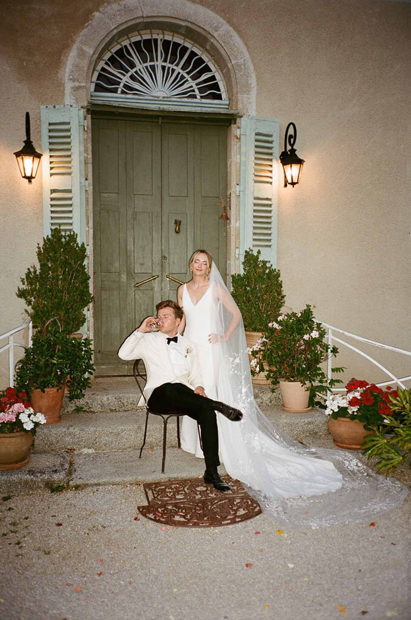 Groom in white dinner jacket seated with drink as bride stands with cathedral lace veil at sage green chateau door