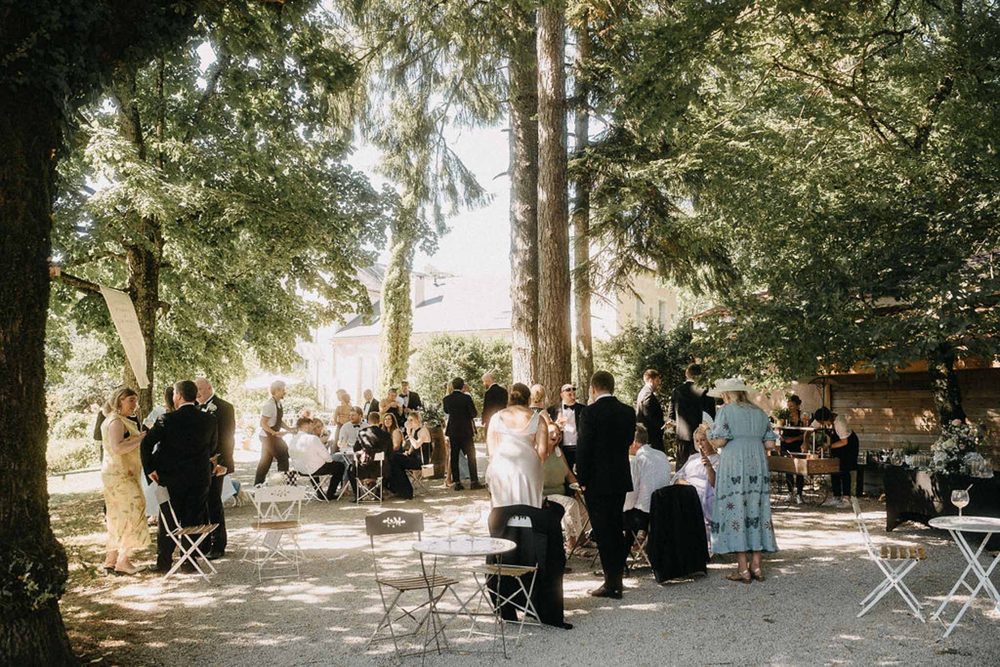 Twenty-eight guests mingle at white bistro tables under tree canopy on gravel terrace at chateau