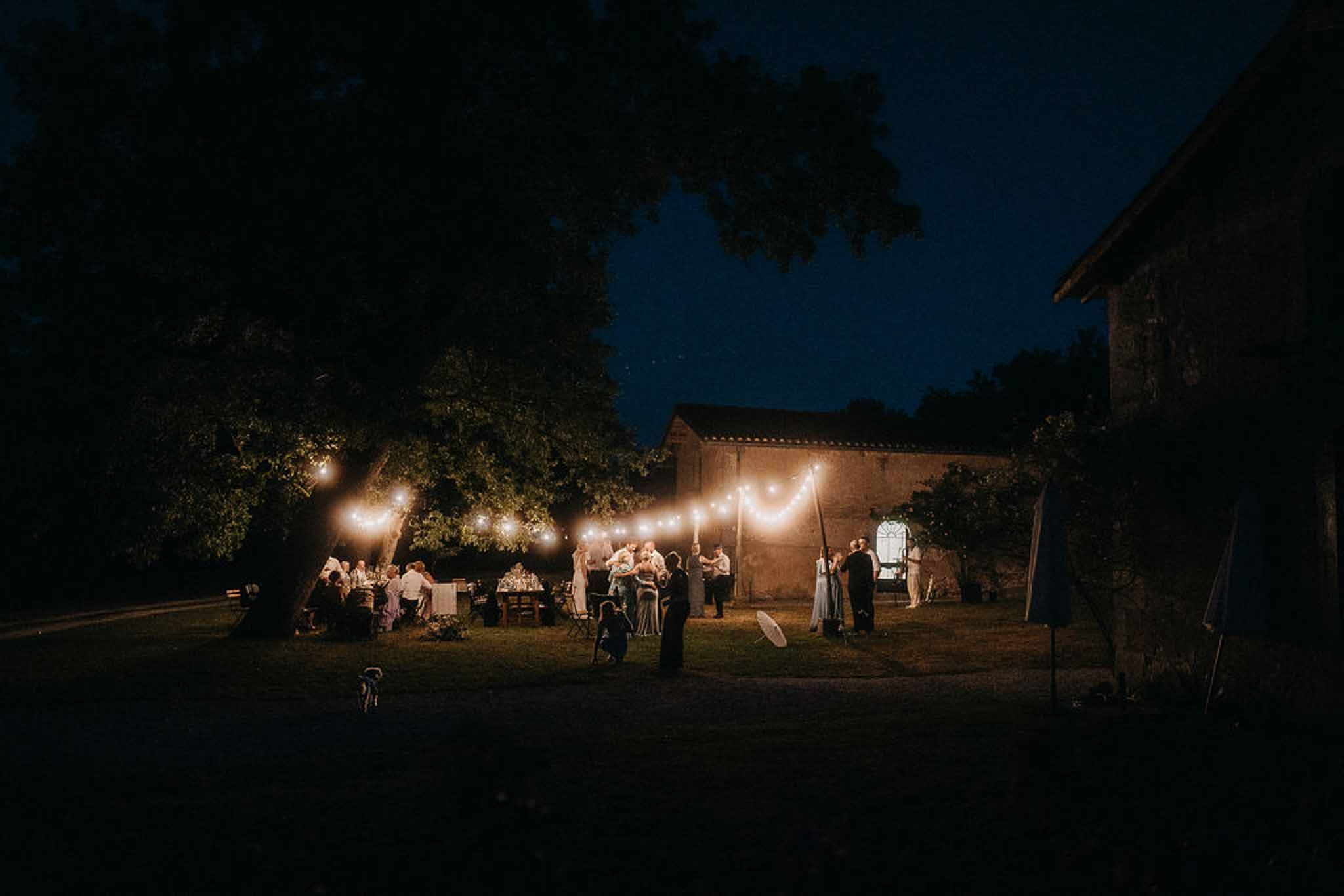 Outdoor evening reception with guests at long tables under festoon lights beside stone farmhouse at night