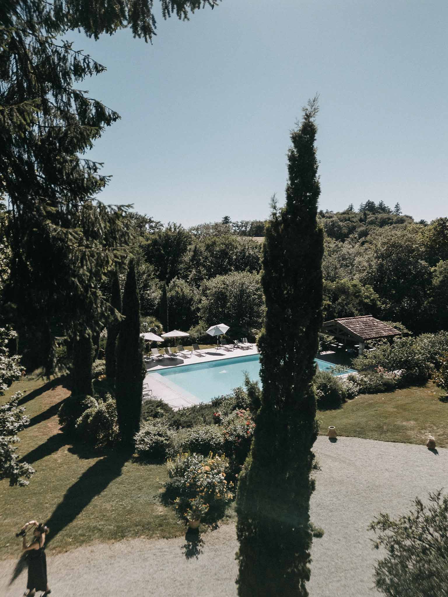 Elevated view of rectangular pool with white parasols, terracotta-roofed pool house, and cypress trees