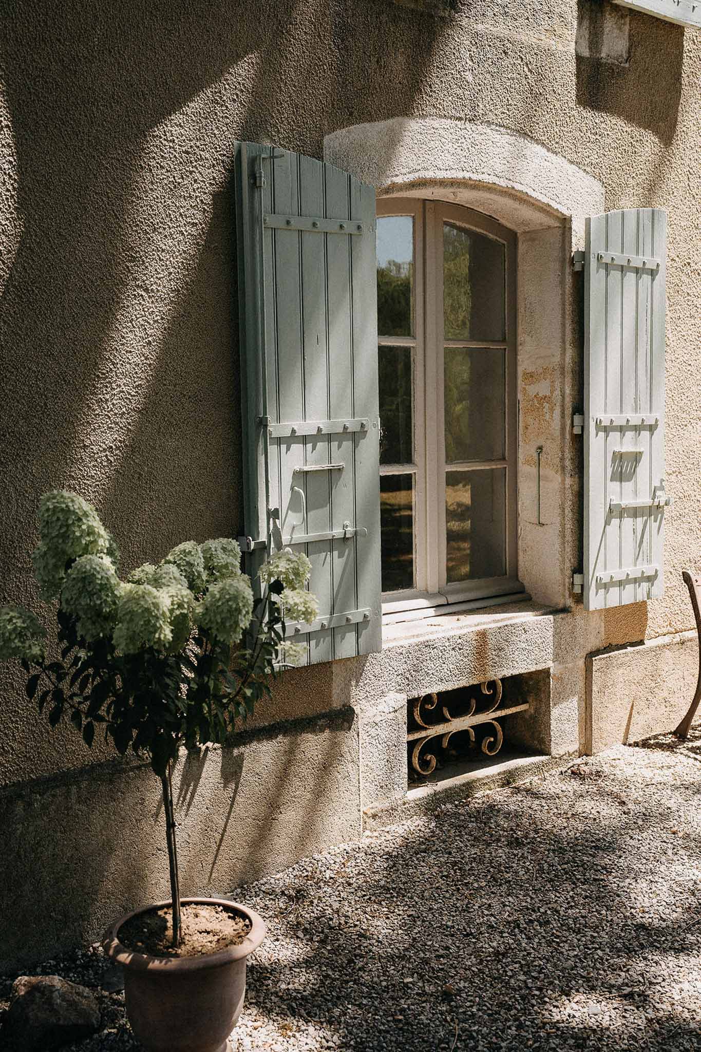 Arched window with sage green shutters on warm beige facade potted white hydrangea standard on gravel at French country pr...
