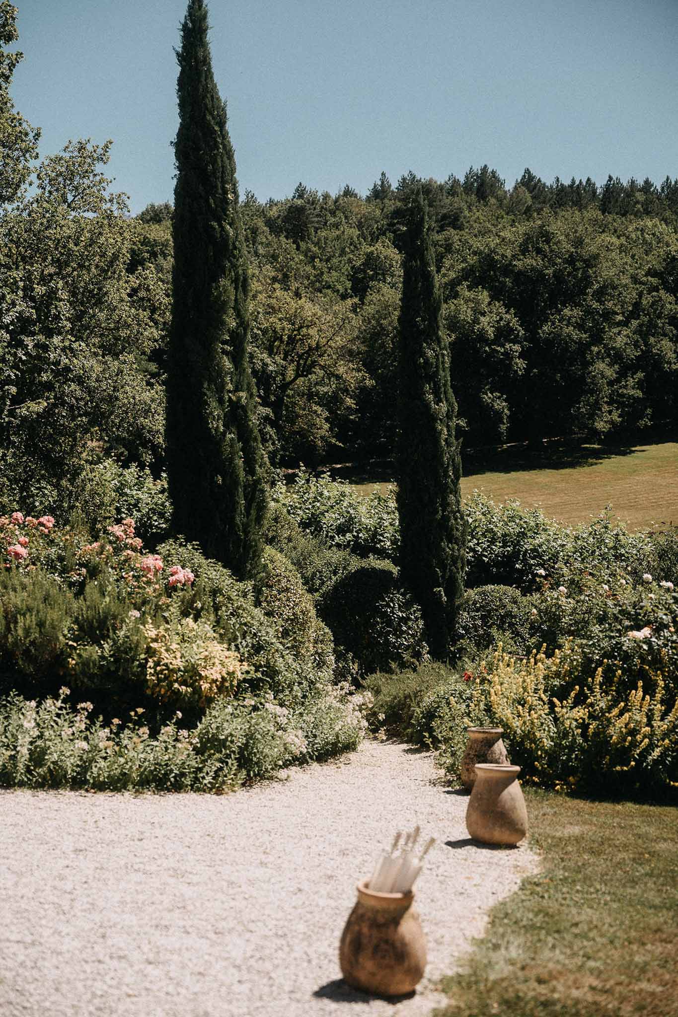 Gravel garden path with pink roses, cypress trees, and terracotta urns holding white parasols