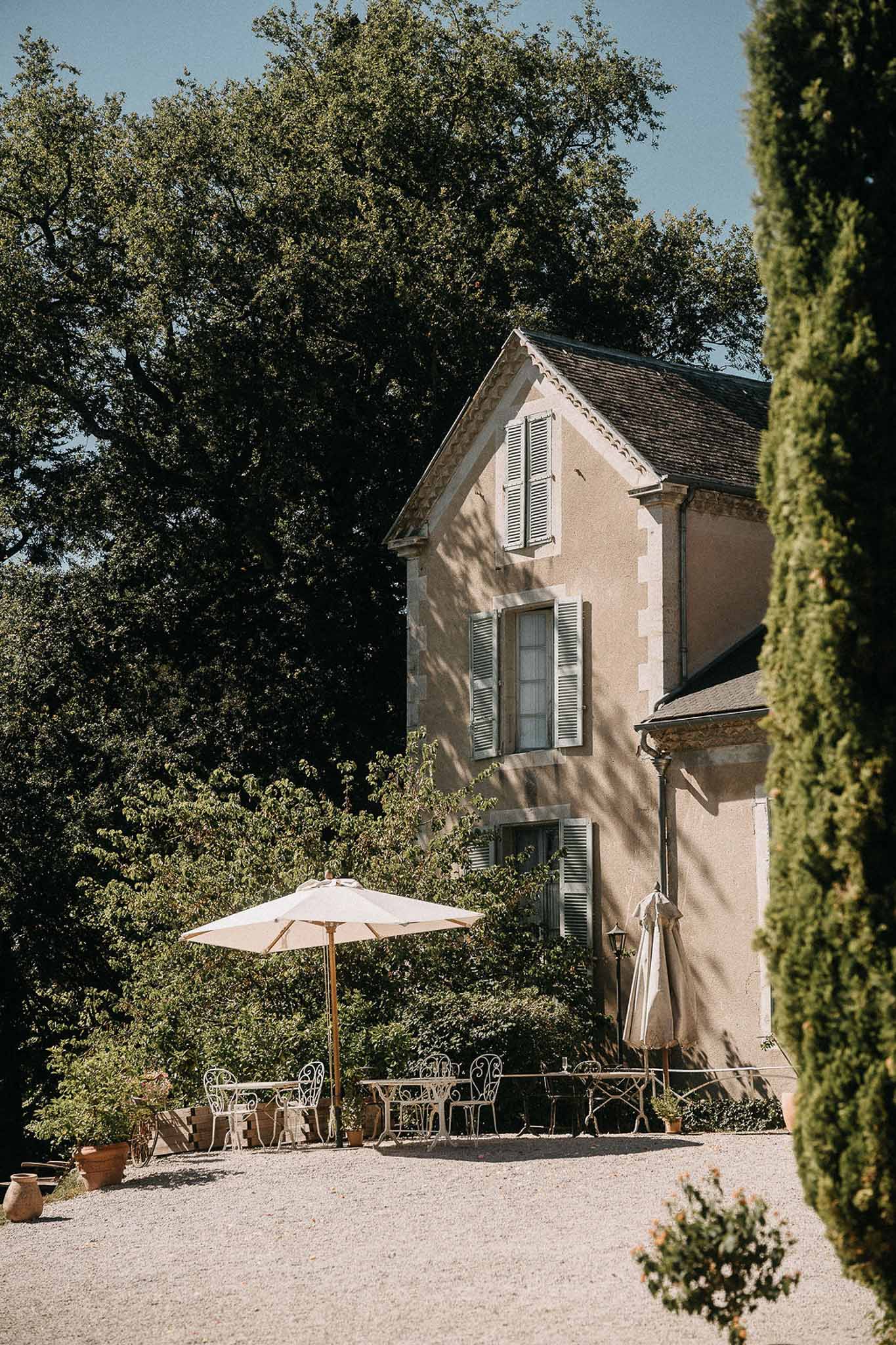 French country manor with sage shutters, terracotta roof, gravel courtyard, bistro tables, and cream umbrellas