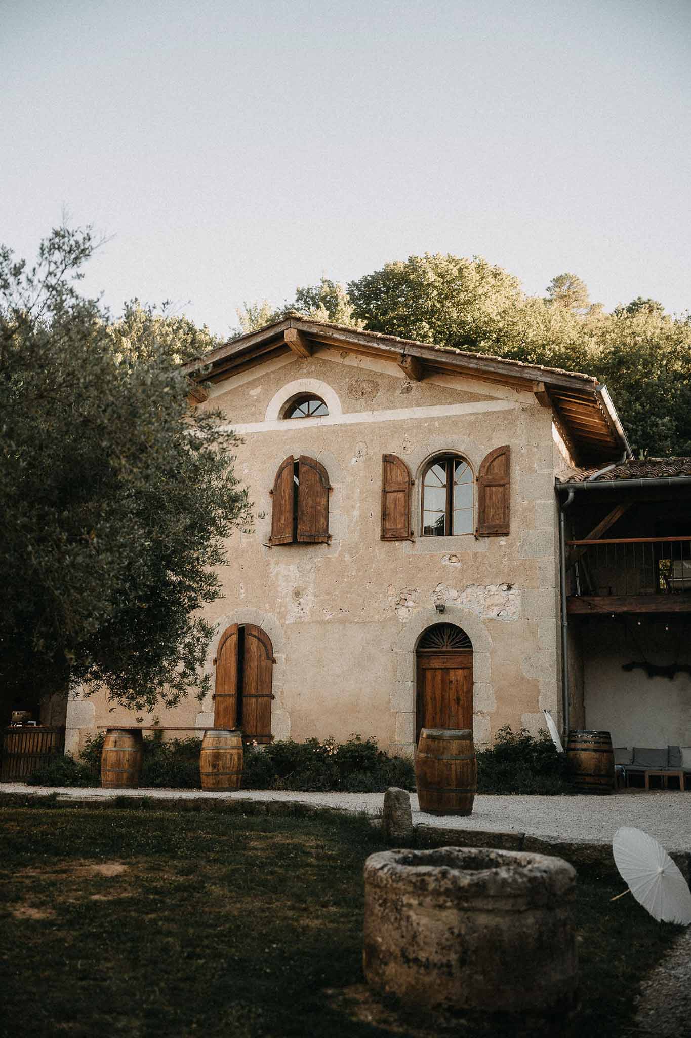 Rustic French stone farmhouse with terracotta roof, oak wine barrels along facade, and stone well in foreground
