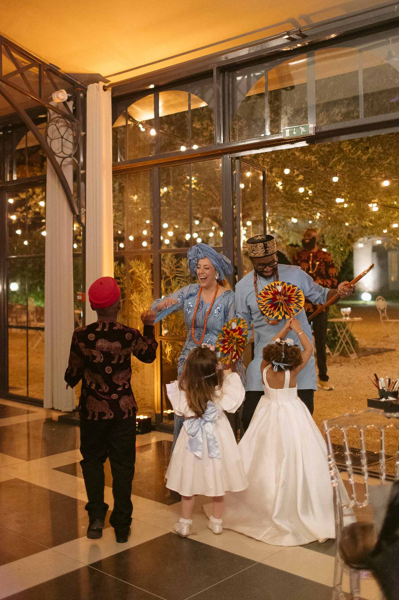 Couple in blue Nigerian aso-oke with flower girls in white dancing in orangery with checkered floor