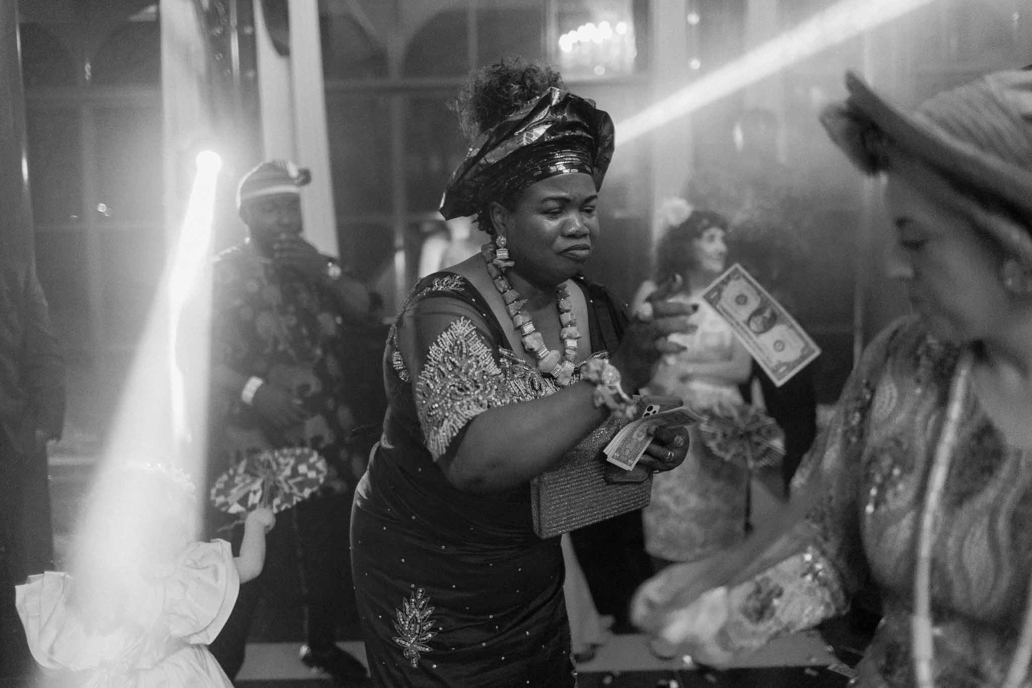 Black and white of guests in traditional dress during money-spraying celebration with dramatic light beams in ballroom