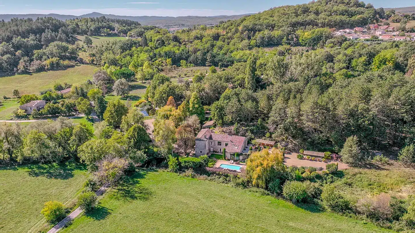 Aerial view of stone country house with pool, gravel courtyard, and wooded valley landscape
