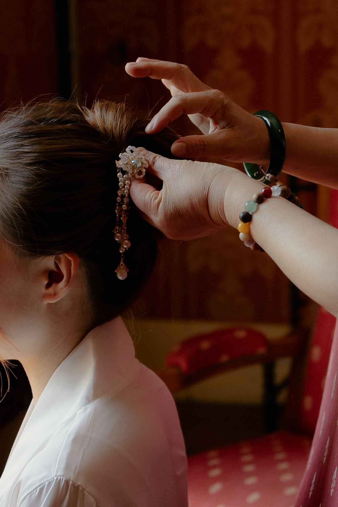 Hands placing pearl and gold chain hair accessory into bride updo in richly decorated chateau room