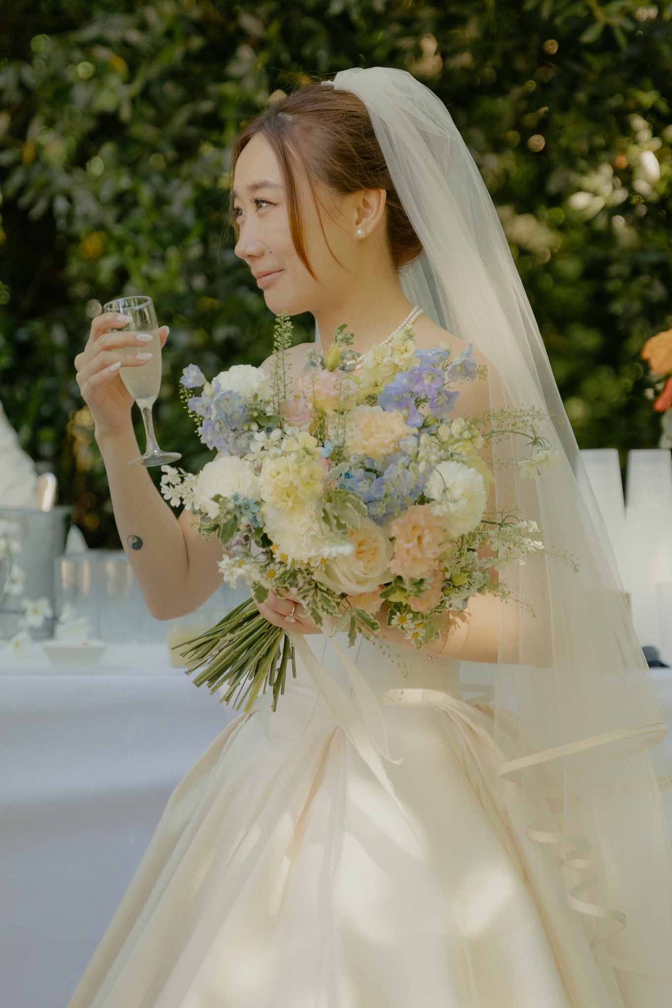 Bride in champagne ballgown with pearl necklace holding pastel bouquet and champagne flute outdoors