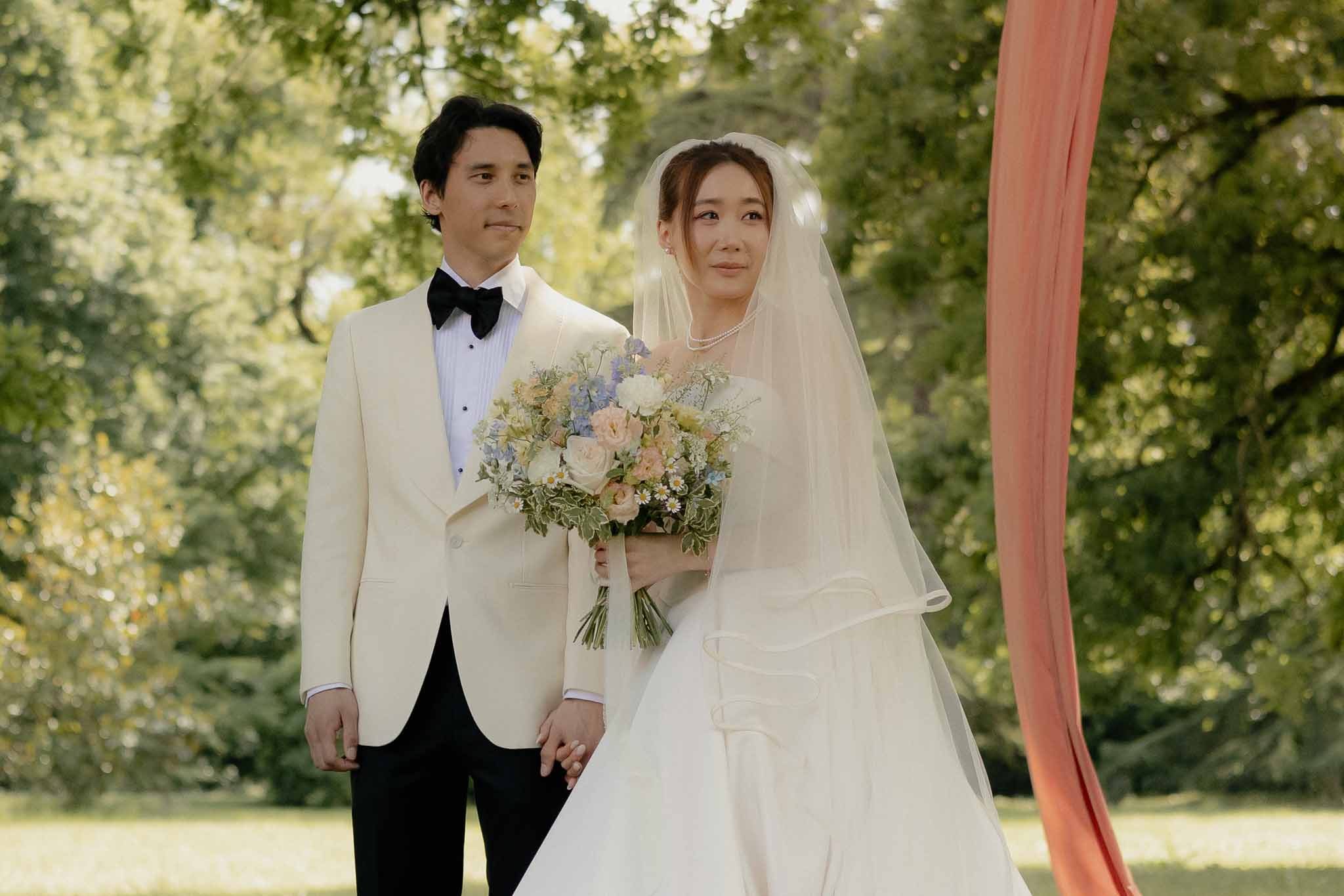 Couple at garden ceremony with blush rose and blue delphinium bouquet and coral fabric arch element