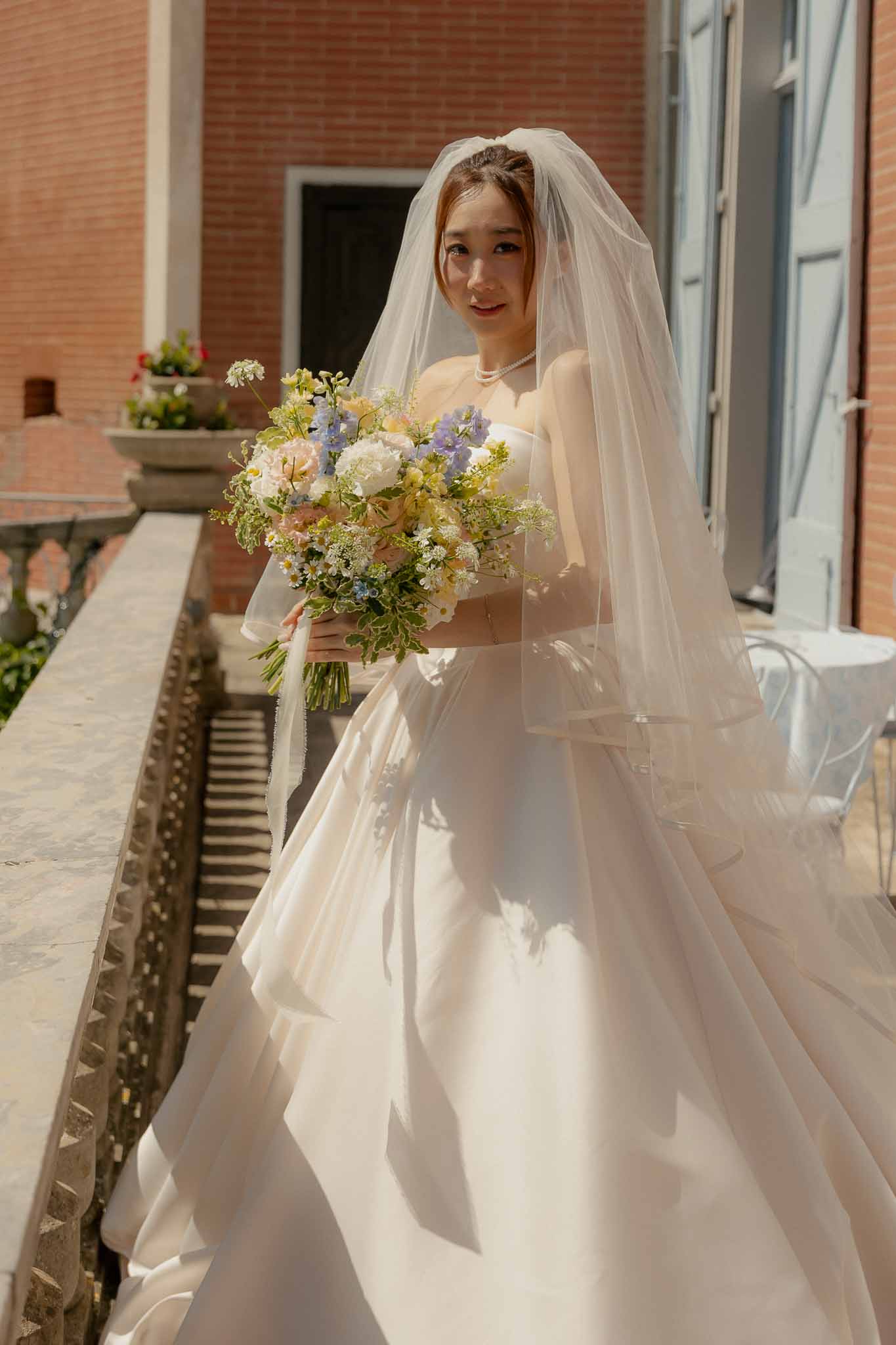 Bride on stone staircase holding garden bouquet of peach roses, white peonies, and blue delphinium