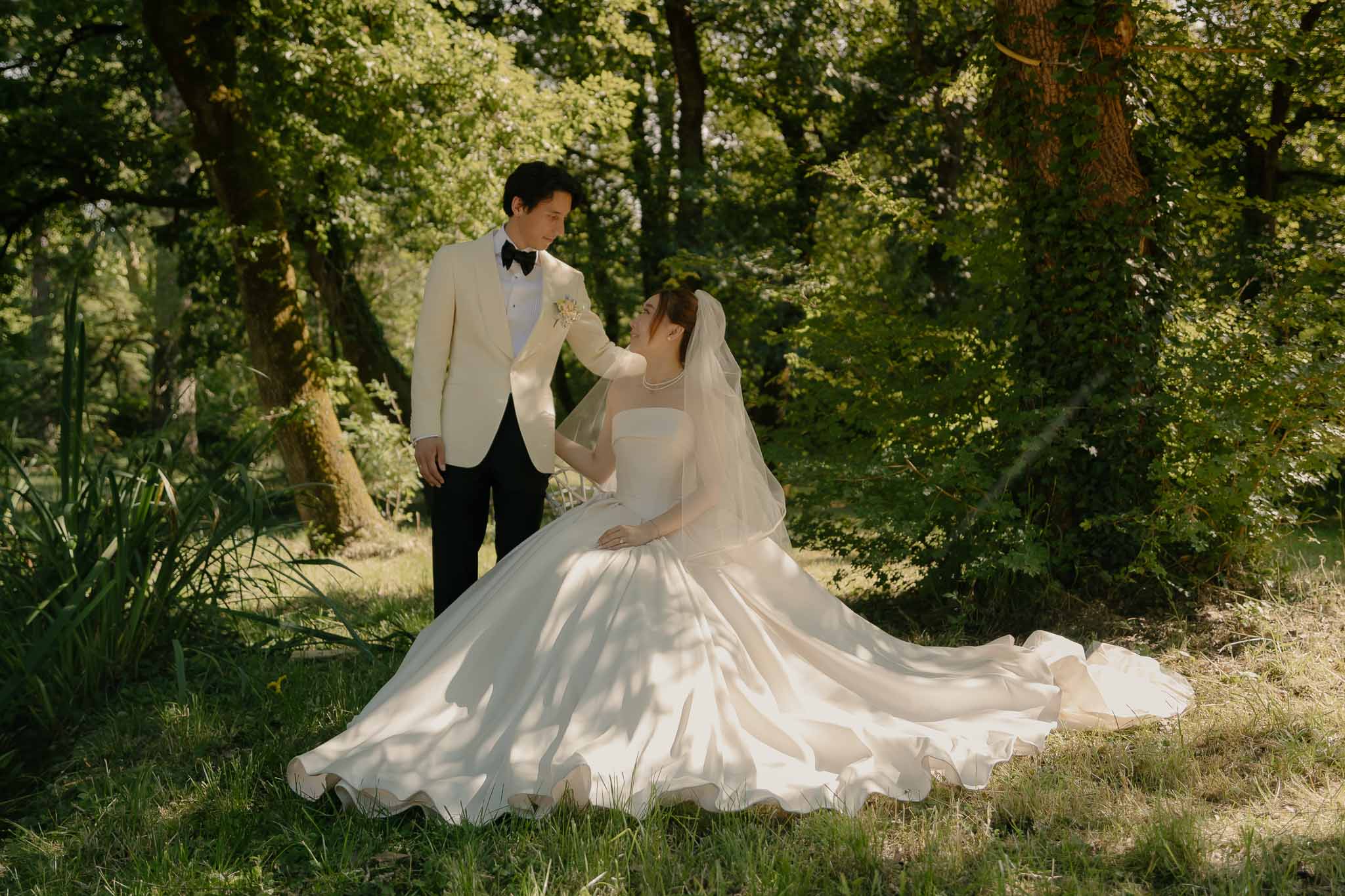Bride seated in spread ivory ballgown with groom standing beside her in wooded garden setting