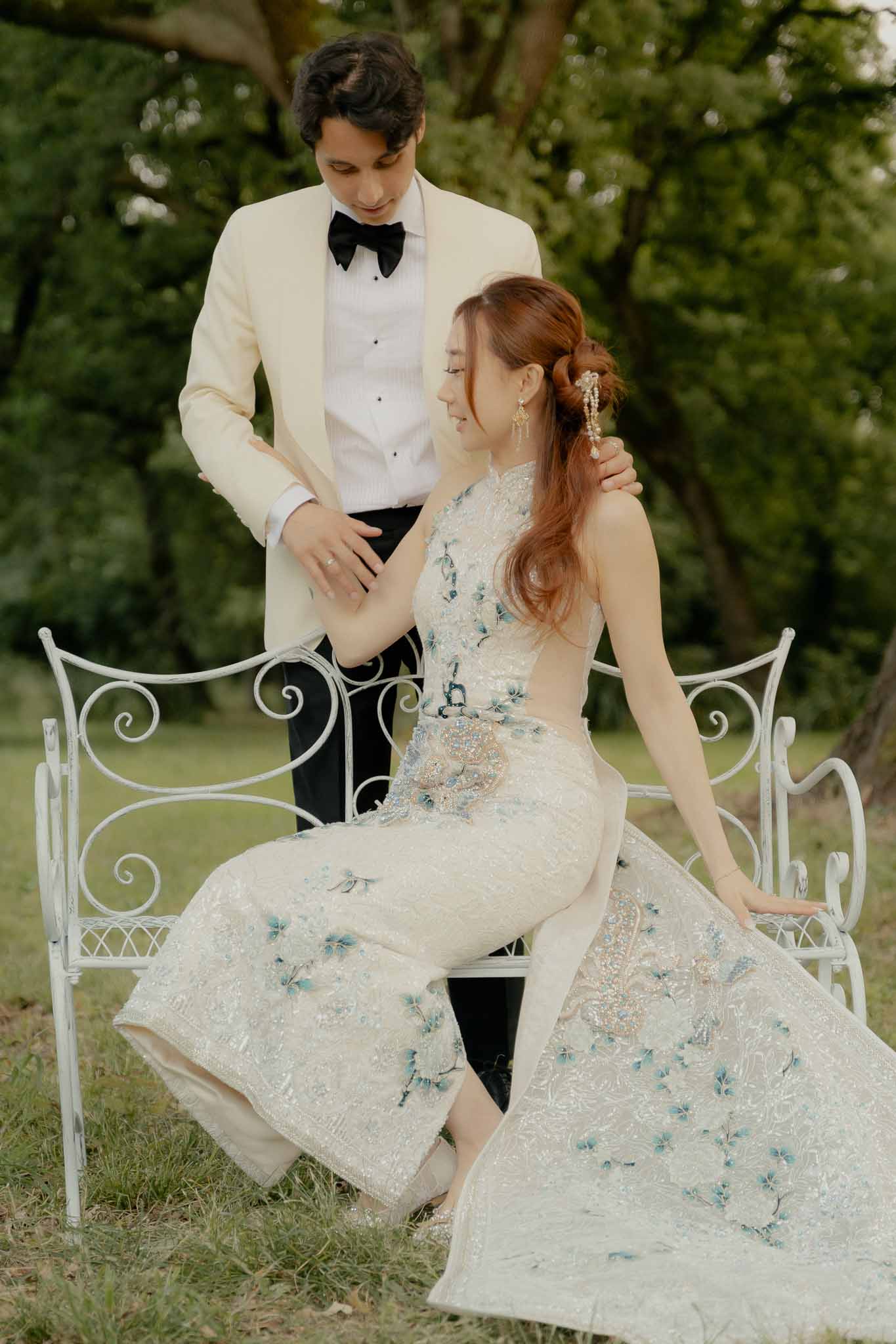 Bride in embellished ivory gown with blue floral embroidery seated on garden bench with groom in cream dinner jacket