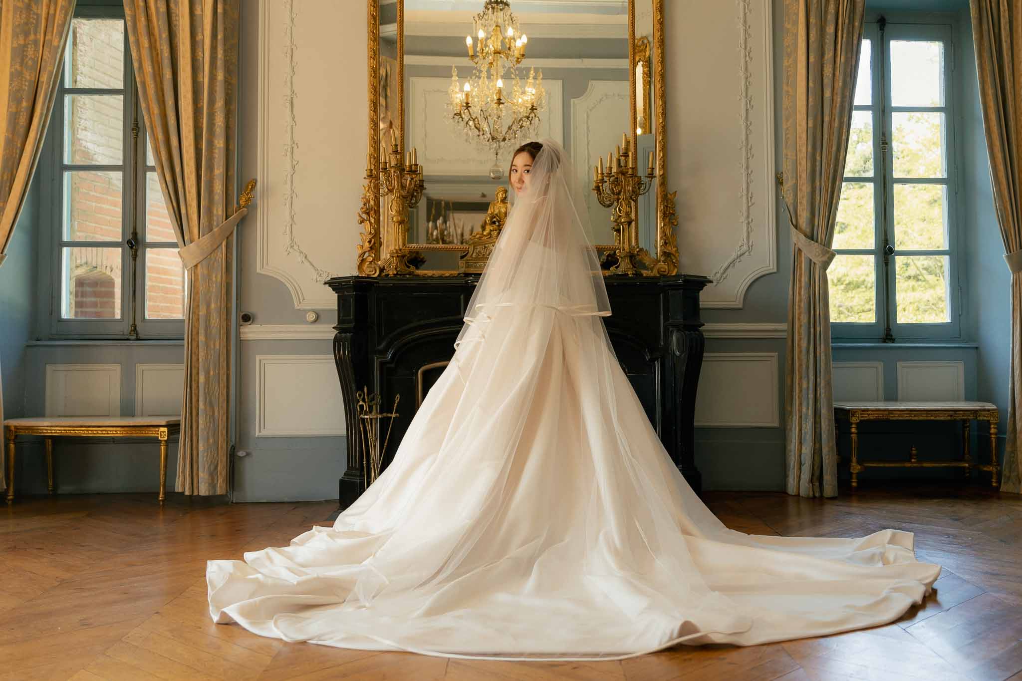 Bride from behind in ivory ballgown with cathedral train and veil in blue-grey chateau salon with gilt mirror