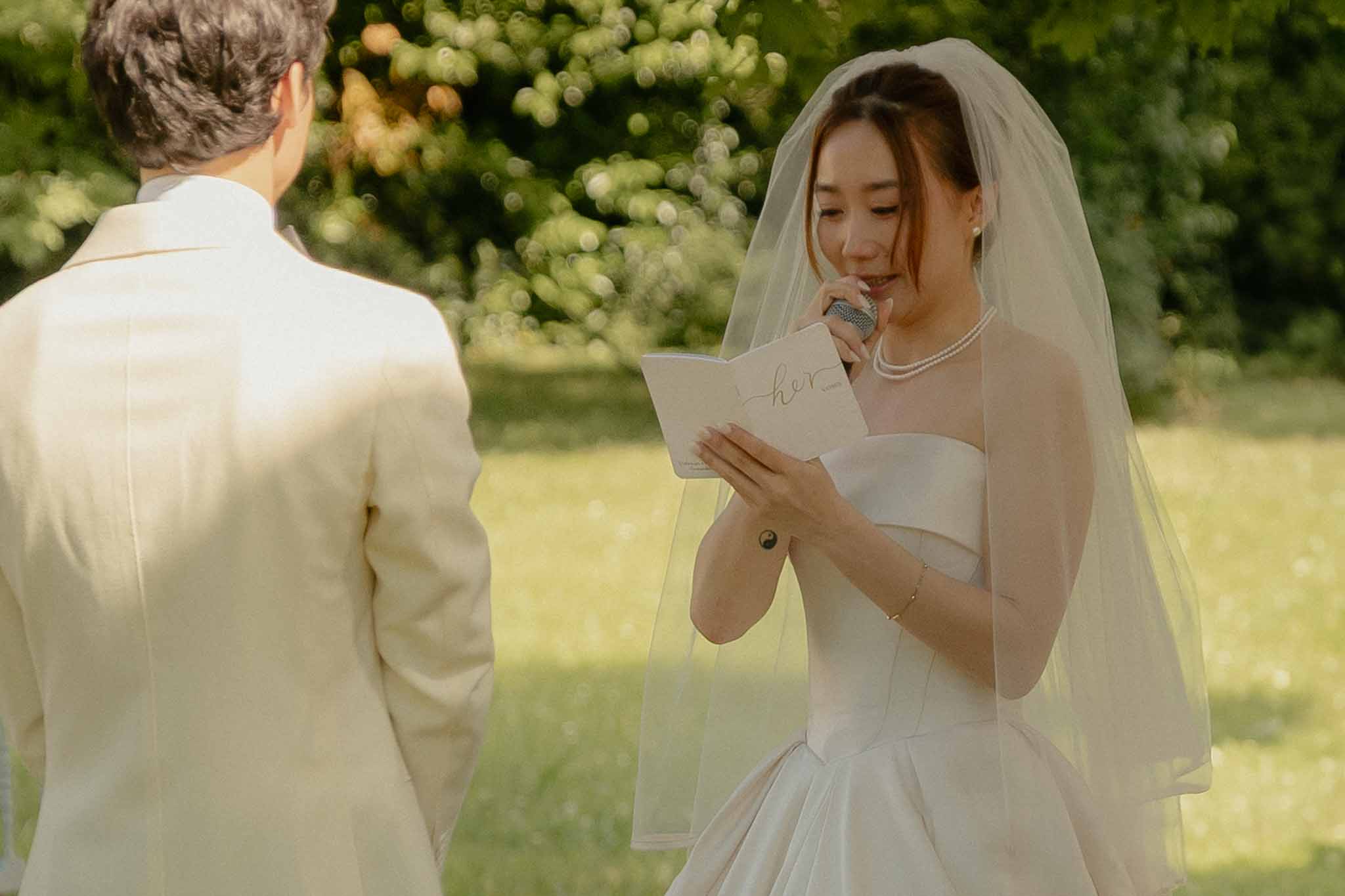 Bride reading personal vows from booklet with microphone during outdoor garden ceremony